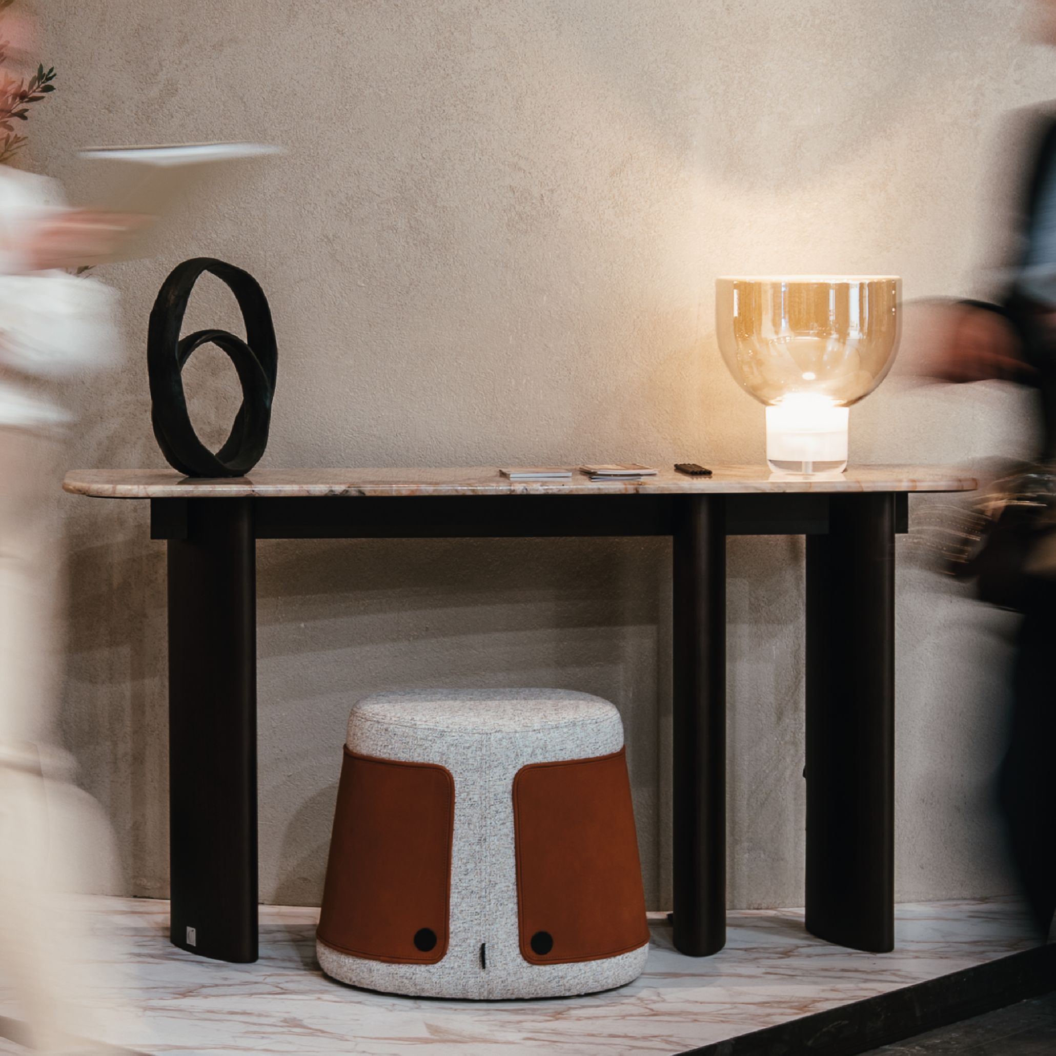 A marble‑topped Dolomites Console stands against a textured microconcrete wall, styled with a black sculptural object, a warm glass lamp, and small decorative pieces. A organic stool sits below while two blurred figures pass by, emphasizing the console’s architectural presence shaped by luxury furniture, enriched by design details, and connected to expressive living room furniture compositions.