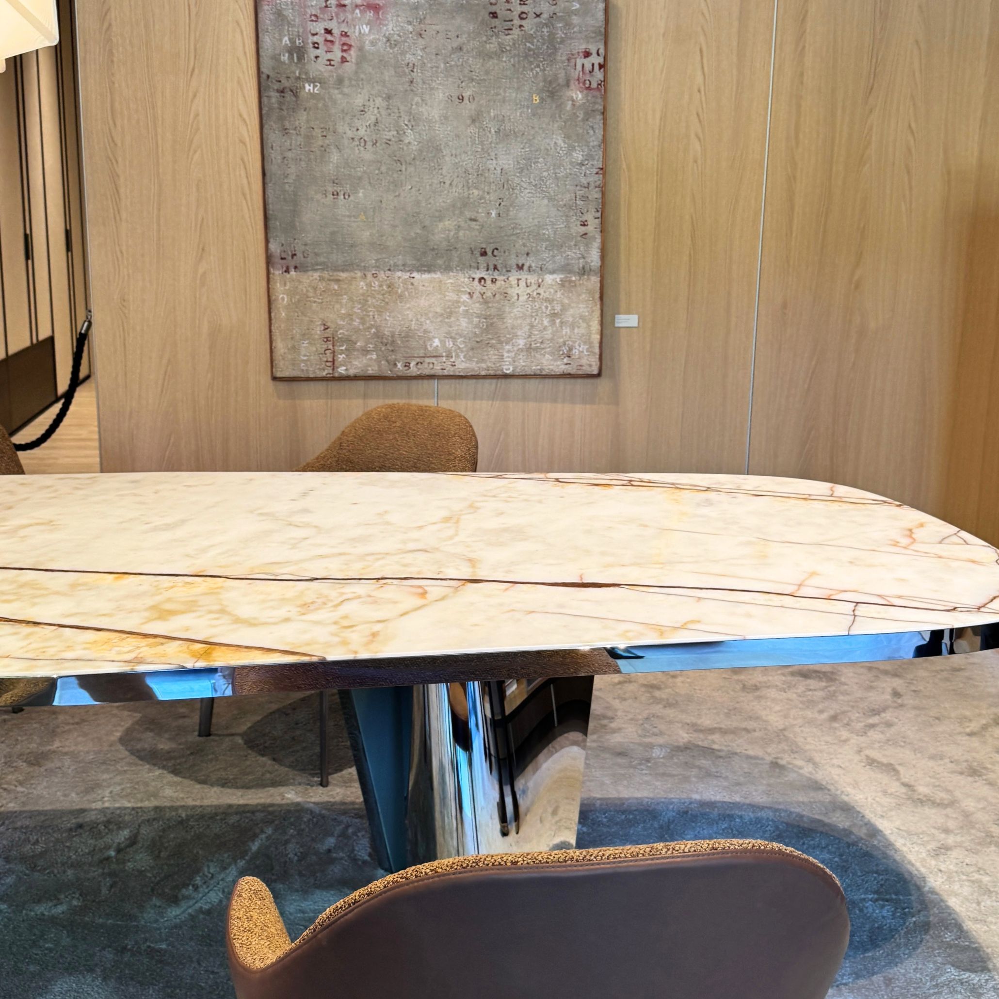 A polished marble table with soft beige and brown veining anchors a warm interior framed by wooden wall panels and an abstract textured artwork. Upholstered brown chairs surround the table, highlighting the trend toward sculptural materials within modern furniture, elevated by luxury home furniture, and guided by atmospheric interior design inspiration.