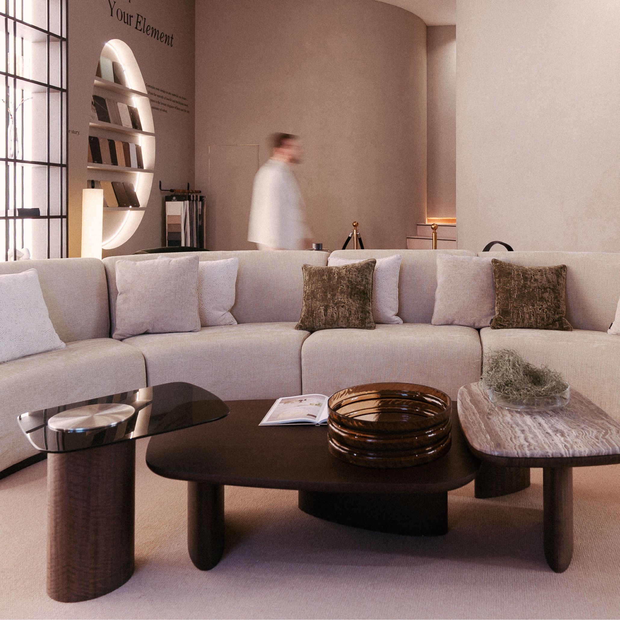A curved beige sectional sofa surrounds the Dolomites Coffee Table, composed of three rounded tables in glass, wood, and stone‑like finishes. Soft lighting highlights how the glass top introduces depth and clarity, contrasting with the warmer, solid materials around it. The warm palette and layered textures reflect expressive surfaces within living room furniture, elevated by luxury home furniture, and shaped by atmospheric design details.