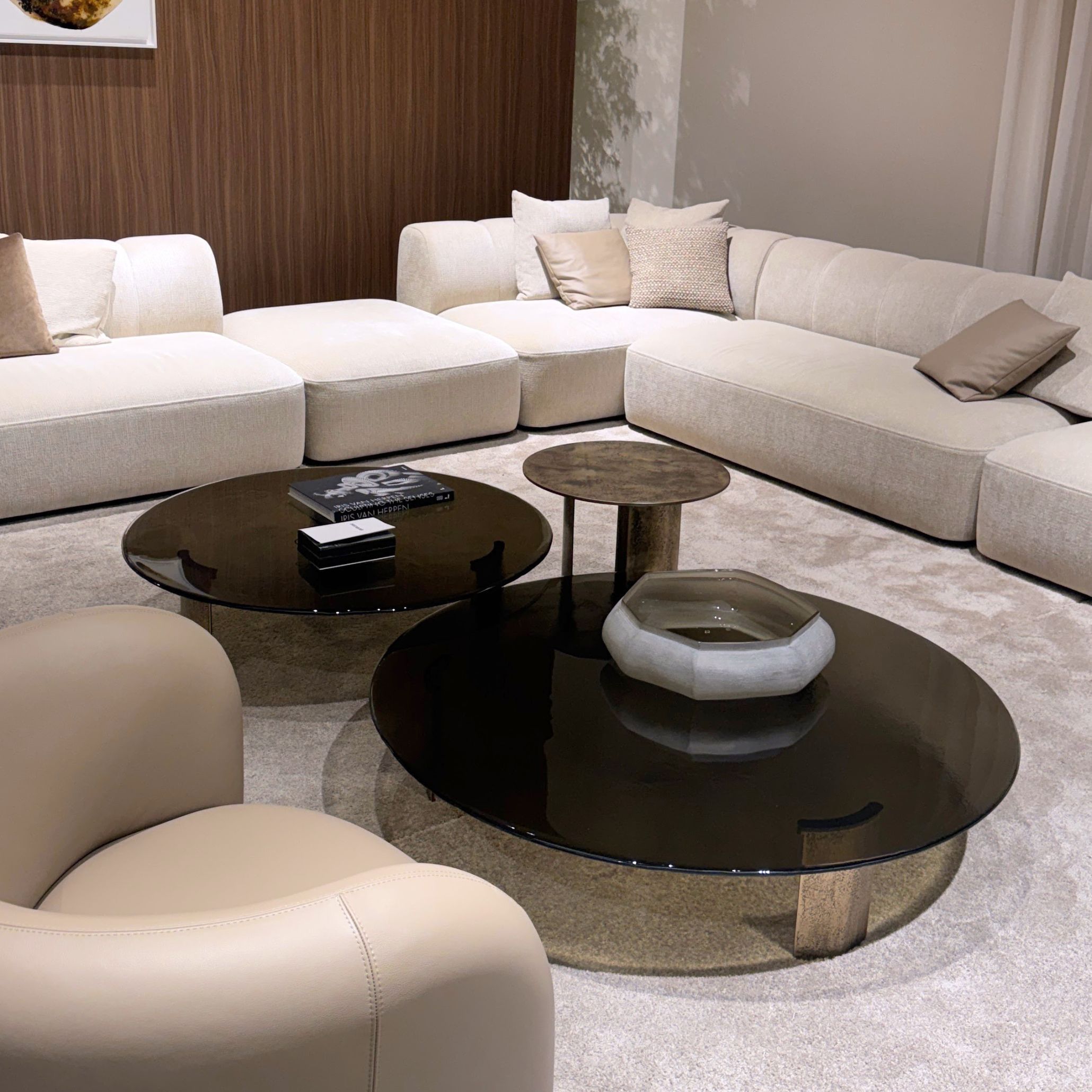 A light beige sectional sofa and armchair frame a trio of rounded coffee tables with dark glass tops and metal legs, styled with books and sculptural decor. The interplay of glass and warm neutrals creates a sense of depth and softness, echoing Salone del Mobile’s focus on transparency within modern furniture, refined luxury furniture, and contemporary interior design inspiration.