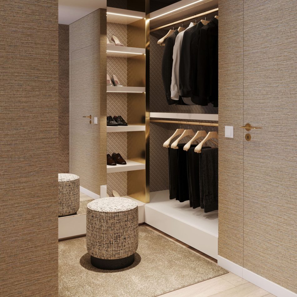 The walk‑in closet features warm beige tones, quilted wall panels, and softly illuminated shelves displaying neatly arranged shoes and clothing. Hanging rods on the right hold jackets and shirts, while a textured upholstered stool sits at the centre of the carpeted floor, adding comfort and balance to the space. This refined environment reflects the elegance of luxury bedroom furniture, shaped by clean contemporary furniture, and elevated through thoughtful design details.
