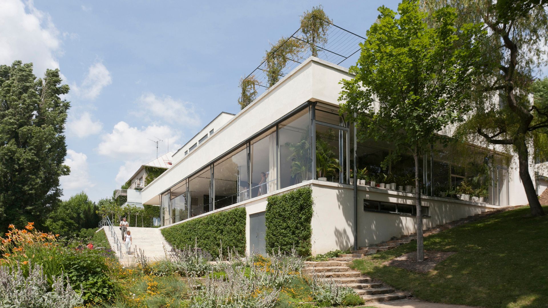 The exterior of Villa Tugendhat reveals its iconic white volumes, expansive glass walls, and floating terraces set within a lush garden. The interplay of open planes, structural clarity, and natural light reflects living room architecture, refined design details, and timeless interior design inspiration that defined Mies van der Rohe’s modernist vision.