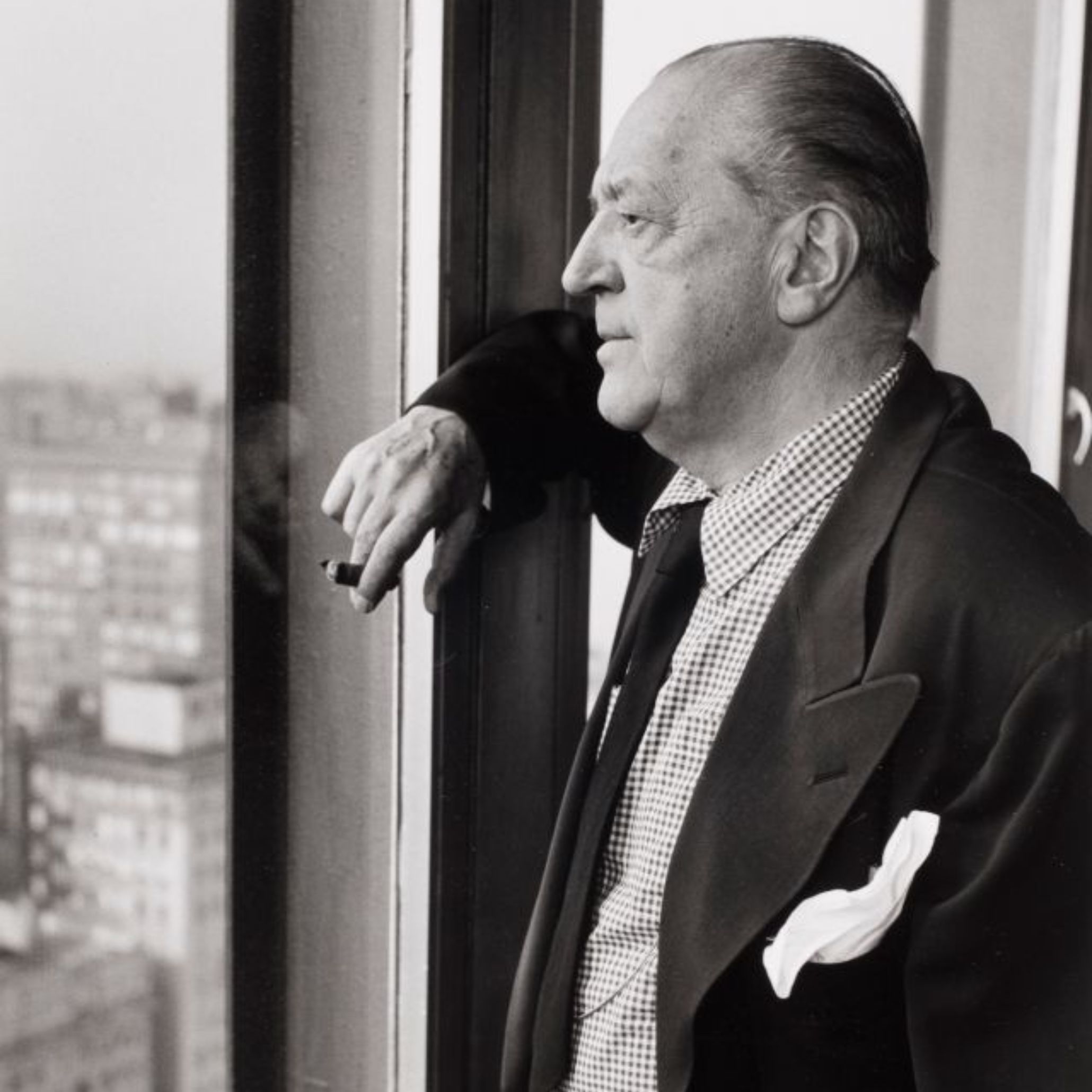 A black‑and‑white portrait of Mies van der Rohe shows him standing by a large window overlooking the city, captured in a moment of quiet reflection. The image conveys the architect’s enduring influence on modern furniture, minimalist design details, and the evolution of contemporary furniture.