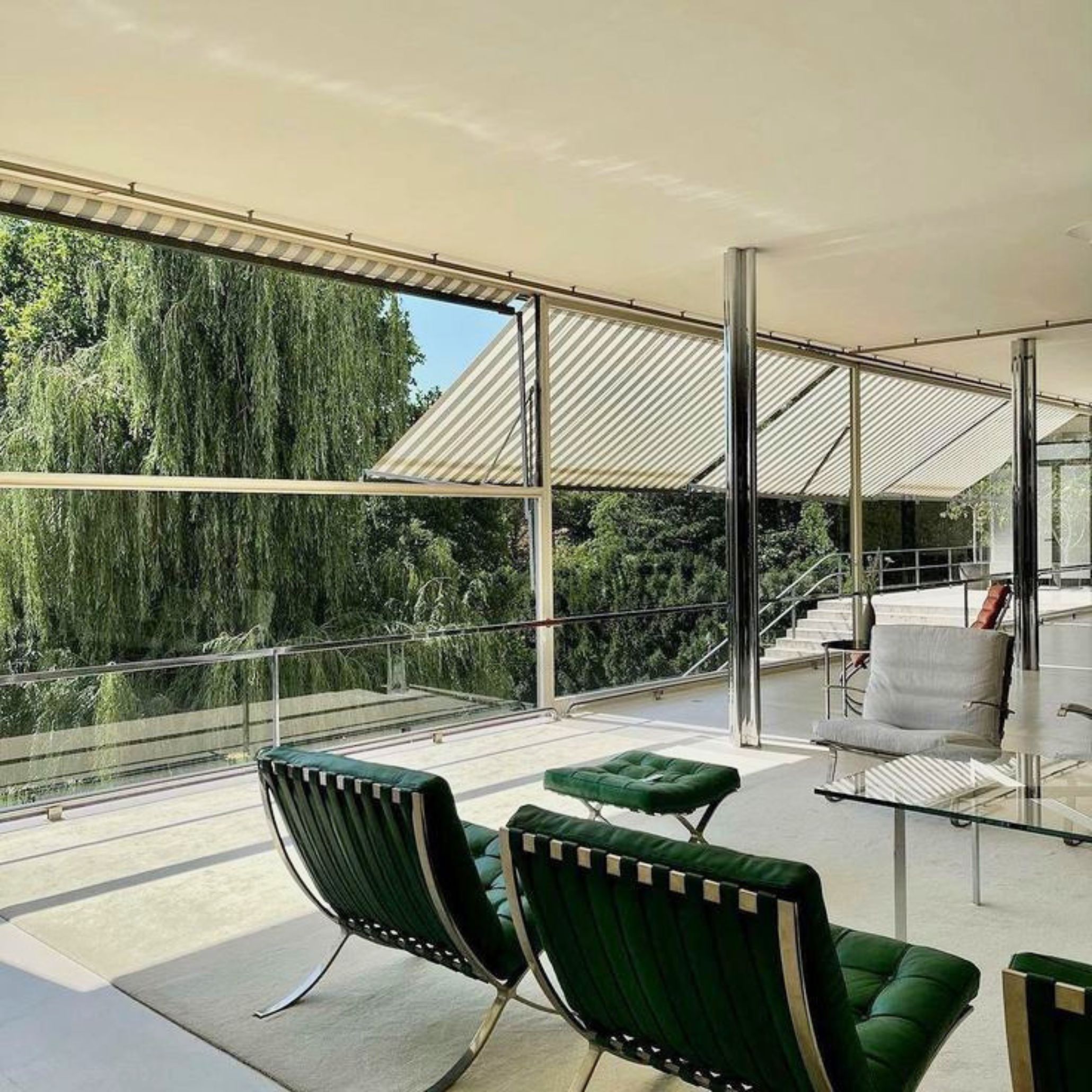 The living area of Villa Tugendhat features floor‑to‑ceiling glass, chrome columns, and minimalist furniture arranged in an open-plan layout overlooking the garden. The space embodies Mies’s modernist principles through luxury home furniture, clean design details, and a fluid sense of living room design.