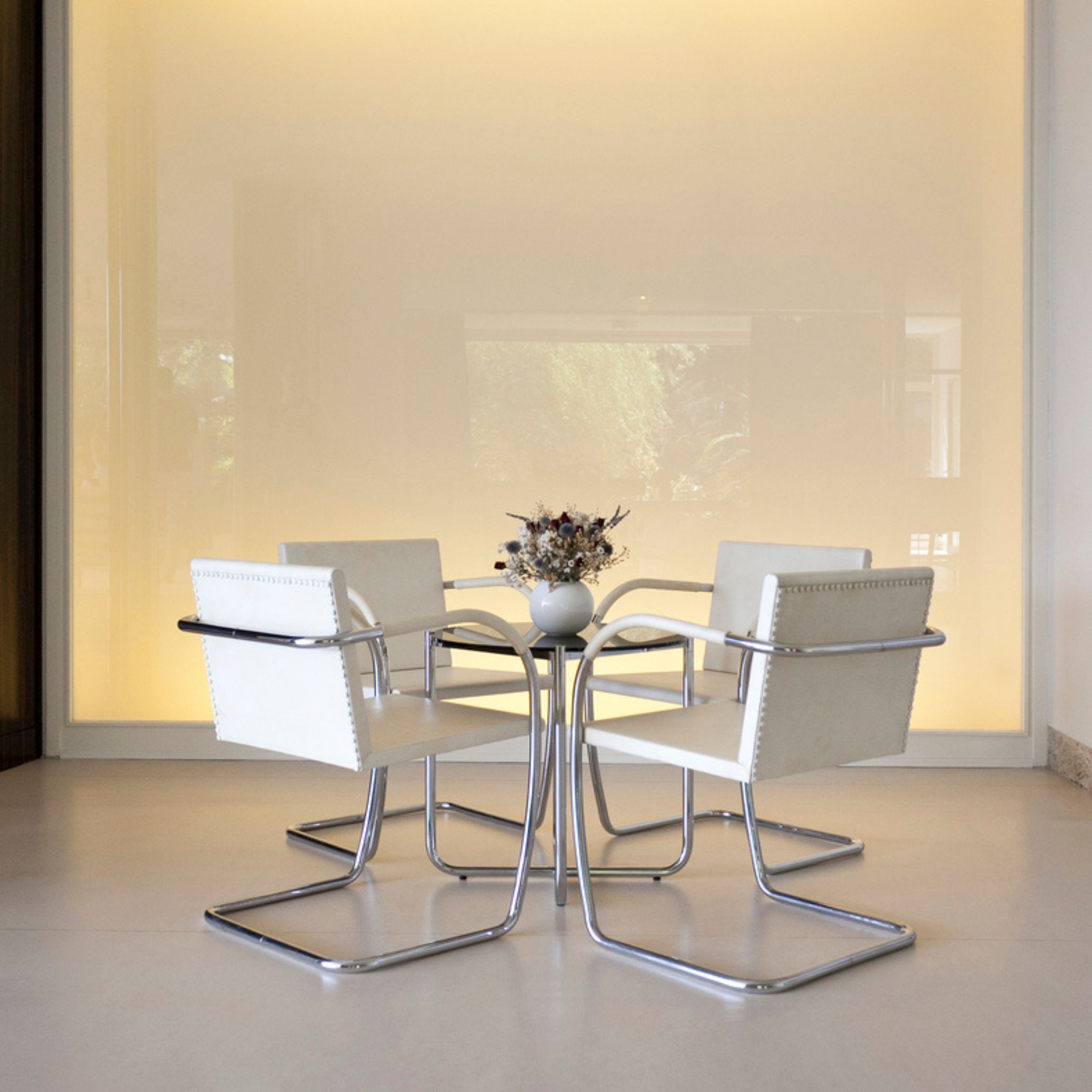 A minimalist dining area inside Villa Tugendhat features a round glass table and white Brno chairs arranged against a softly illuminated wall, expressing Mies van der Rohe’s clarity of form through design details, modern furniture, and serene interior design inspiration.