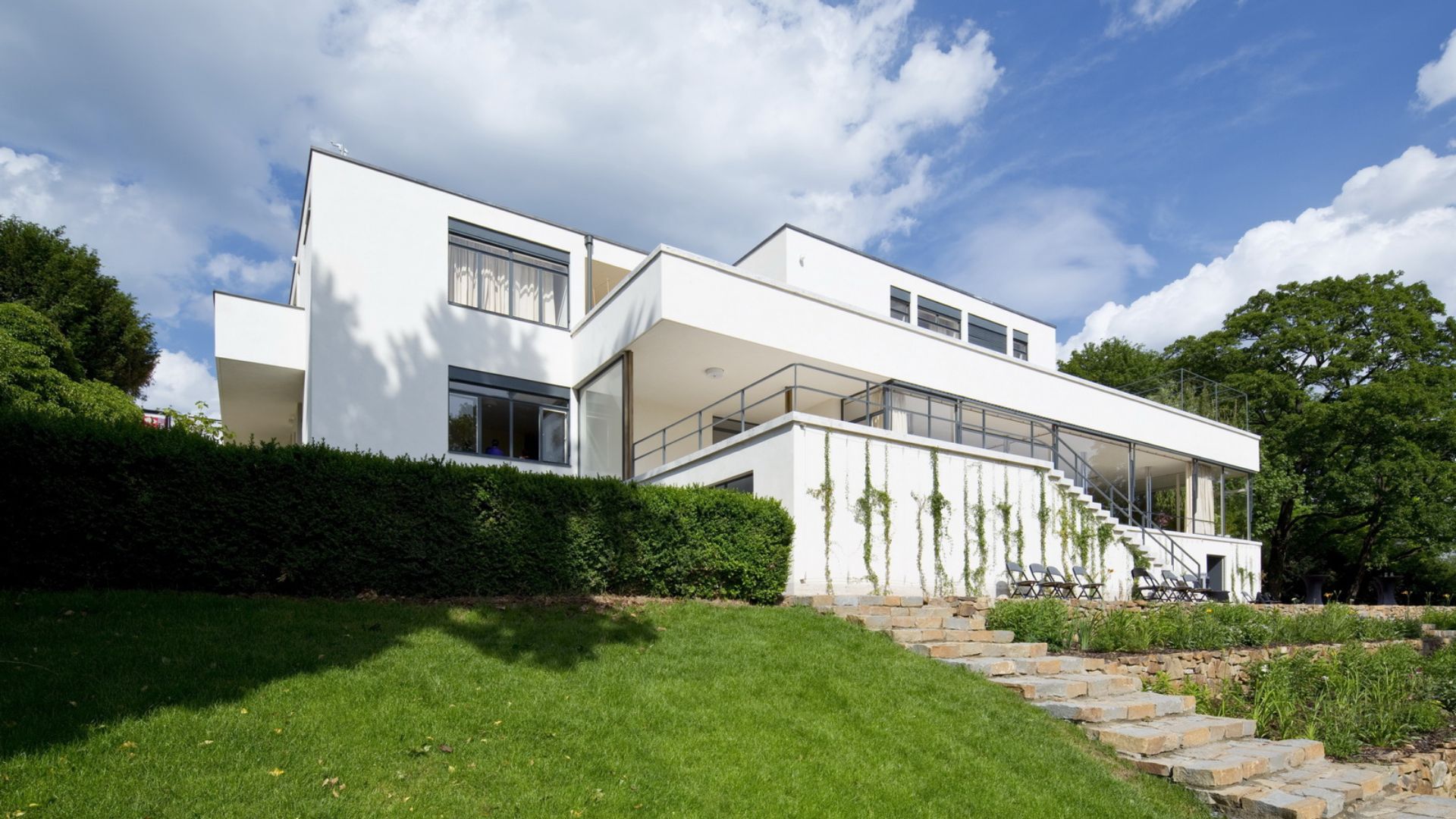 The restored exterior of Villa Tugendhat reveals its clean white volumes, open terraces, and geometric lines set against a landscaped garden. The clarity of form and renewed materials highlight the villa’s modernist character through refined design details, timeless living room architecture, and enduring interior design inspiration.