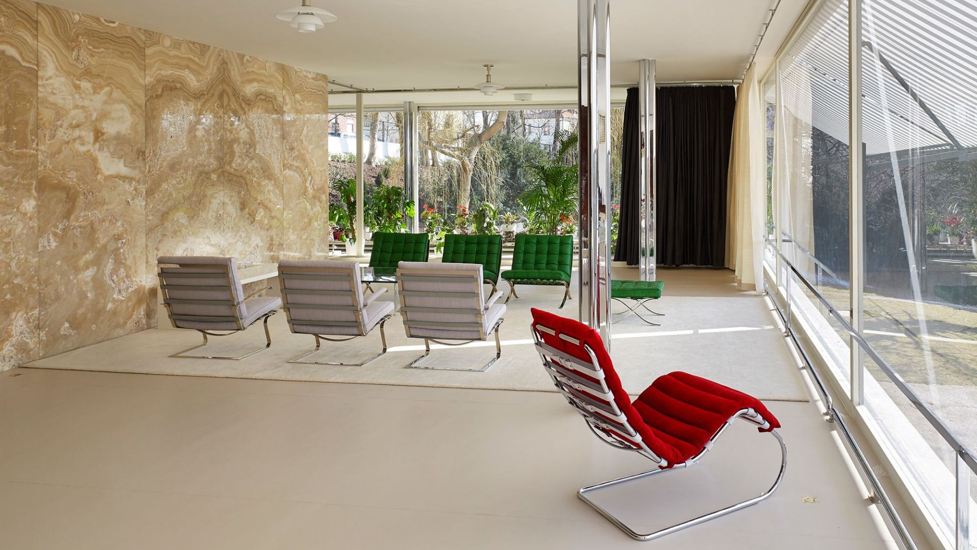 A modernist living space inside Villa Tugendhat features chrome‑framed chairs in muted tones, a striking red accent chair, and a polished stone wall illuminated by natural light from the full glass façade. The open-plan composition reflects Mies van der Rohe’s clarity of proportion through refined design details, sculptural modern furniture, and the seamless openness central to living room architecture.