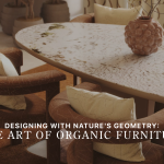 The Stone dining table with soft mineral veining sits surrounded by curved upholstered chairs, illuminated by warm natural light and framed by earthy textures. A sculptural vase with delicate branches anchors the composition, introducing the calm, grounded aesthetic of nature‑led design. The scene reflects expressive organic furniture, the warm tactility of natural interiors, and the sculptural clarity of contemporary design.