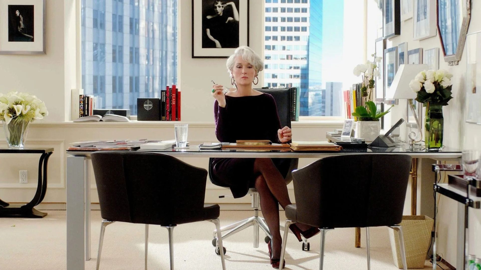 A confident editor sits at a sleek desk surrounded by framed photography, white florals, and expansive windows that flood the workspace with natural light, creating a bright, editorial atmosphere. The balanced composition blends modern office furniture, subtle design details, and refined interior design inspiration, capturing the luminous precision of the Miranda Priestly aesthetic.