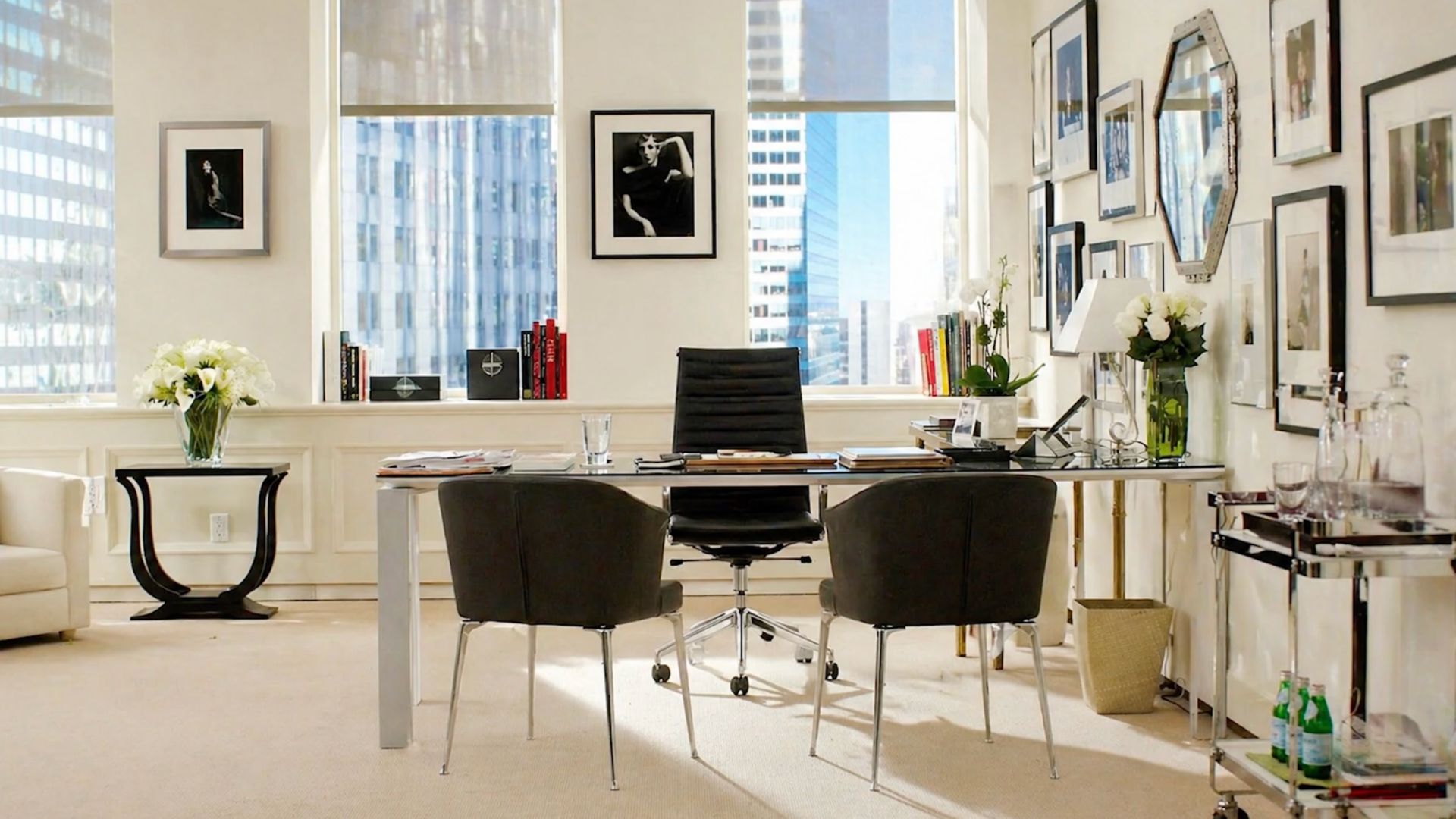 A sleek, monochrome office inspired by Miranda Priestly features a glass desk, black leather chair, and curated wall art arranged with editorial precision. The clean palette, metallic accents, and structured layout reflect modern office furniture, elevated design details, and refined interior design inspiration that echo the quiet authority of the Devil Wears Prada aesthetic.
