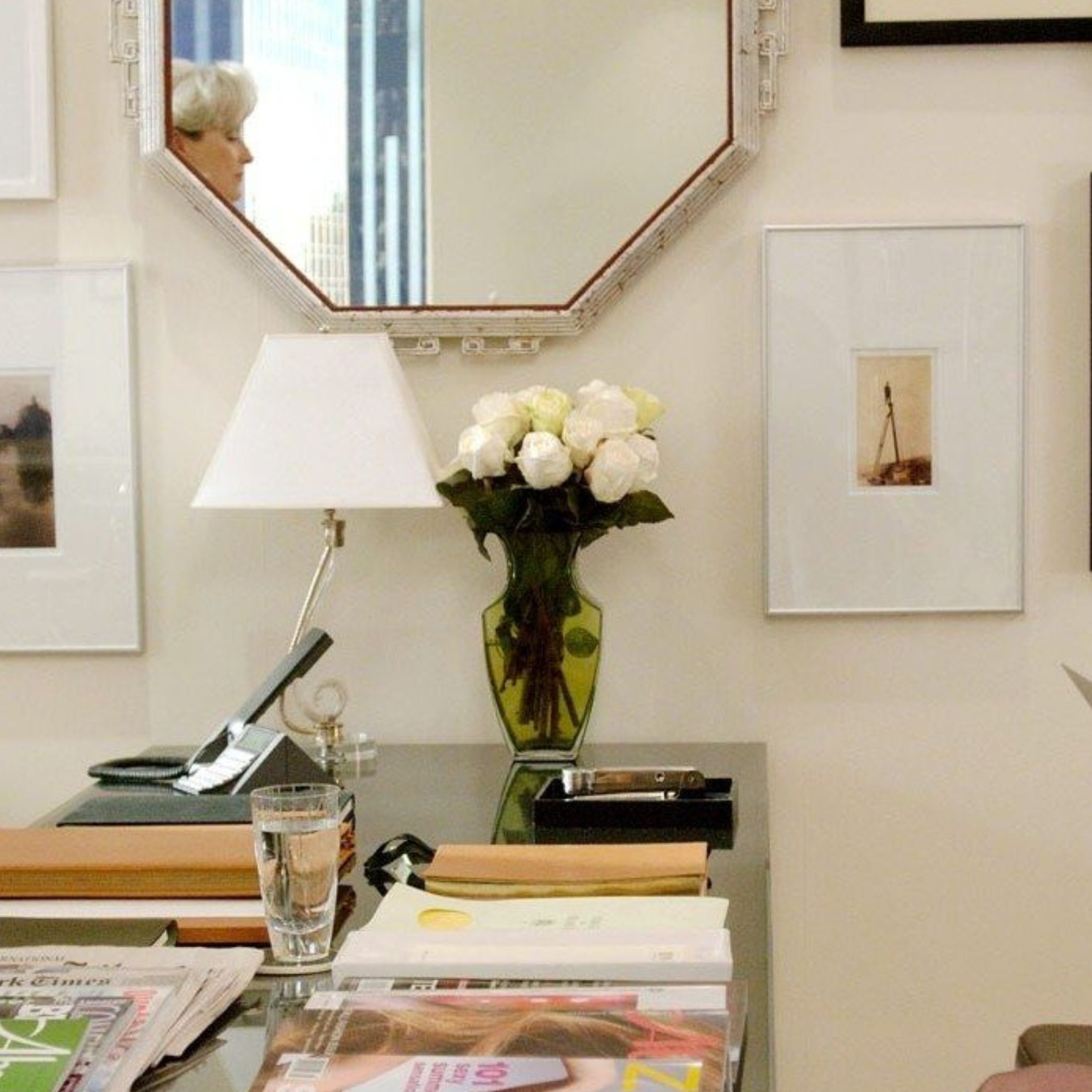 A neatly styled desk features white roses in a green vase, paired with curated books, framed artwork, and a reflective mirror that adds depth to the workspace. The composition blends modern office furniture, subtle design details, and refined interior design inspiration to soften the structured Miranda Priestly aesthetic.