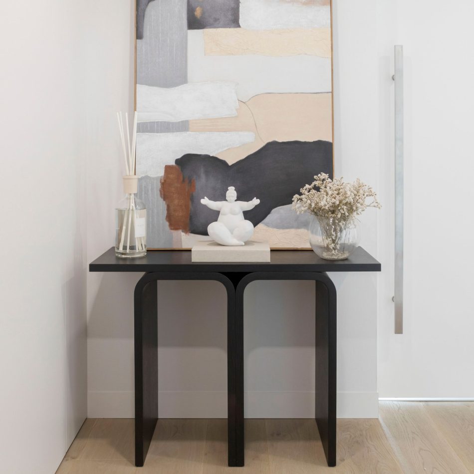 The Antónia Console appears in a black finish, its curved legs forming a sculptural silhouette beneath a large abstract artwork. Decorative objects, a diffuser, a seated sculpture and a glass vase with dried stems, soften the composition, creating a calm atmosphere. The scene mixes modern furniture design, warm contemporary decor and the quiet balance of minimalist interiors.