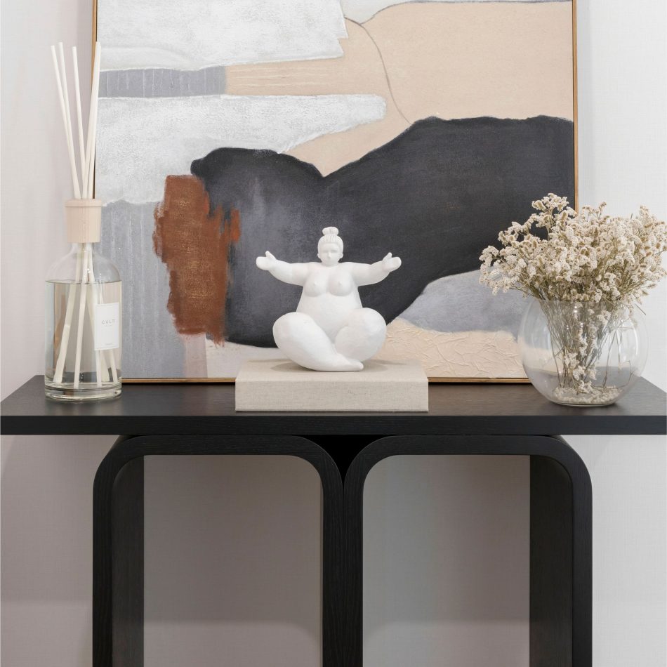 A close view of the Antónia Console reveals its smooth black surface and elegant curved supports, styled with a sculptural figure, a diffuser and a vase of dried flowers. The abstract artwork behind it adds depth and texture, shaping a serene, modern ambiance. The moment reflects sculptural modern furniture design, expressive contemporary decor and understated minimalist interiors.