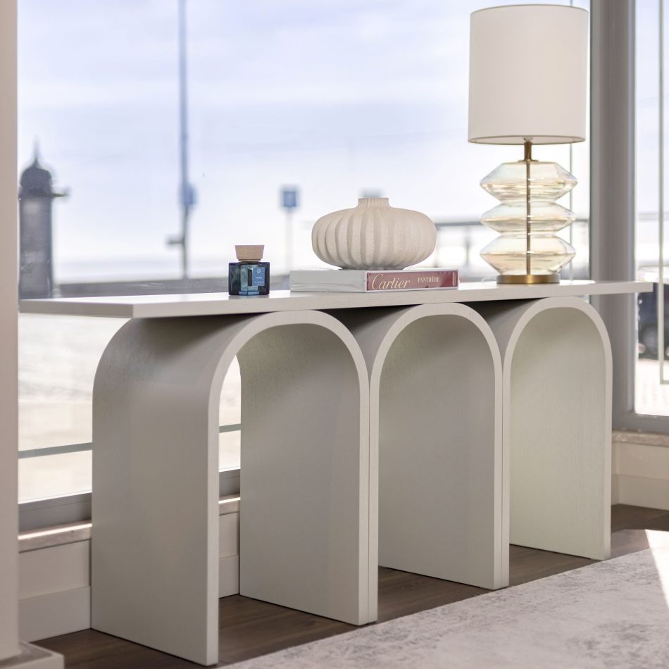 The neutral Antónia Console stands near a large window, its three arches forming a sculptural silhouette softened by natural daylight. A ribbed ceramic vase, a design book and a glass lamp create a calm, curated arrangement that enhances the room’s airy, coastal ambiance. The scene reflects modern furniture design, warm contemporary decor and the serene balance of minimalist interiors.