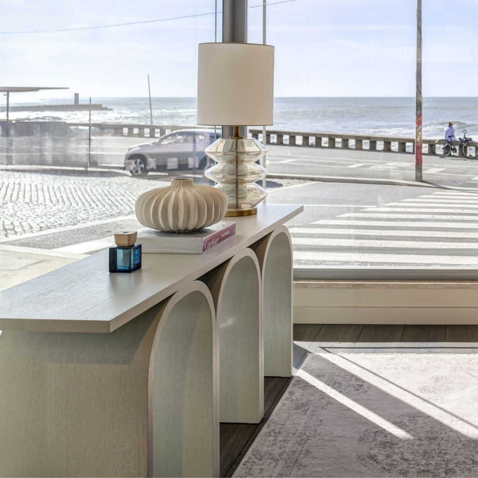 The Antónia Console appears in a bright, open corner overlooking a sunlit seaside street, its arched structure grounding the space with quiet elegance. A sculptural ceramic vase, a blue glass container and a layered glass lamp create a soft, harmonious composition that complements the coastal light. The moment reflects sculptural modern furniture design, expressive contemporary decor and the understated clarity of minimalist interiors.