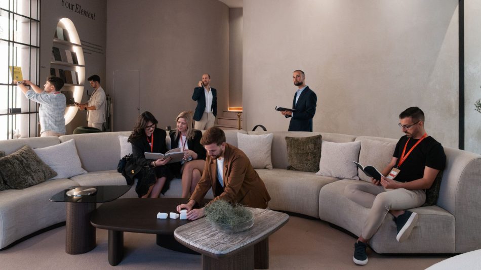 Members of the Stylish Club team move through the warm, minimalist stand, some reading on the curved beige sofa while others stand browsing objects and materials around the space. The layered coffee tables, soft neutrals, and gentle lighting frame a candid, distracted moment that reflects the human side of luxury furniture, shaped by expressive living room furniture, and enriched by contemporary interior design inspiration.