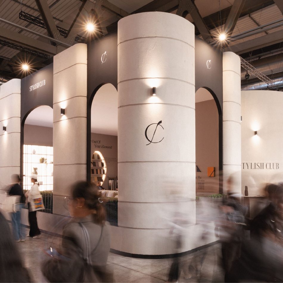 The Stylish Club stand at Salone del Mobile 2026 features tall cylindrical columns, arched openings, and warm beige surfaces accented with black lighting details. Visitors walk past the architectural structure, highlighting the brand’s sculptural identity within luxury furniture, shaped by expressive design details, and aligned with contemporary interior design inspiration.