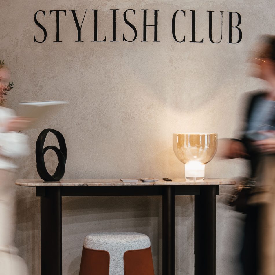 A marble‑topped console with a black sculptural object and warm glass lamp stands against a textured wall marked “Stylish Club,” while two blurred figures pass by. The scene captures the stand’s refined materiality within modern furniture, enriched by luxury home furniture, and guided by atmospheric living room furniture compositions.