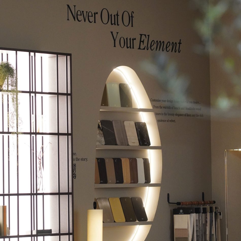 An illuminated oval niche displays material samples, woods, metals, and lacquers finishes, beneath the phrase “Never Out Of Your Element,” with fabric swatches and greenery nearby. The setup reflects Stylish Club’s focus on customization within high end furniture, elevated by tactile design details, and connected to contemporary interior design inspiration.