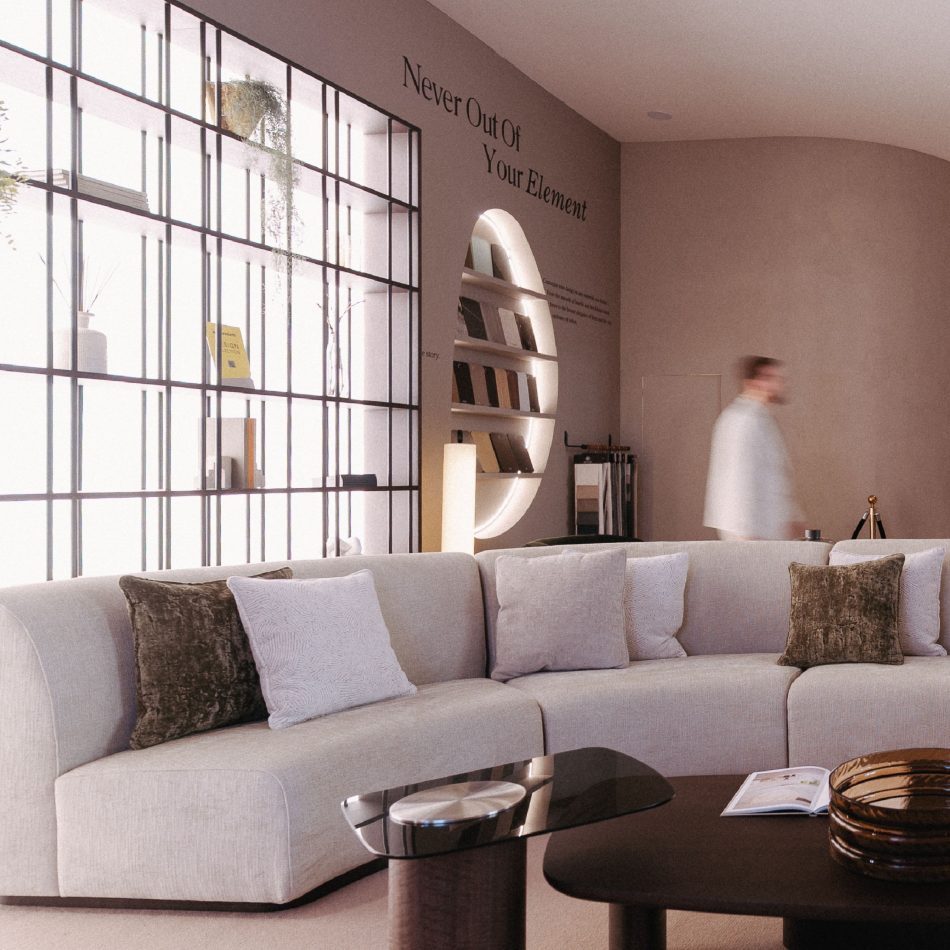 A curved Stone Sofa in soft beige anchors the space, layered with textured cushions in white and olive tones. A coffee table set with a glass bowl and an open magazine sit in front, while a windowed shelving wall and the phrase “Never Out Of Your Element” frame the warm, contemporary atmosphere.