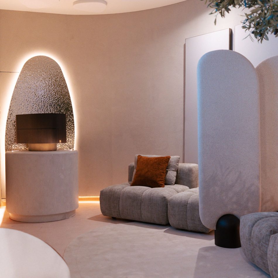 A quiet private area features a neutral antónia sofa with a rust‑toned cushion, a cylindrical side table with a textured lamp, and a curved partition with soft, rounded edges. A glowing oval wall panel adds warmth, shaping an intimate corner defined by bedroom furniture and minimalist high end furniture sensibility.