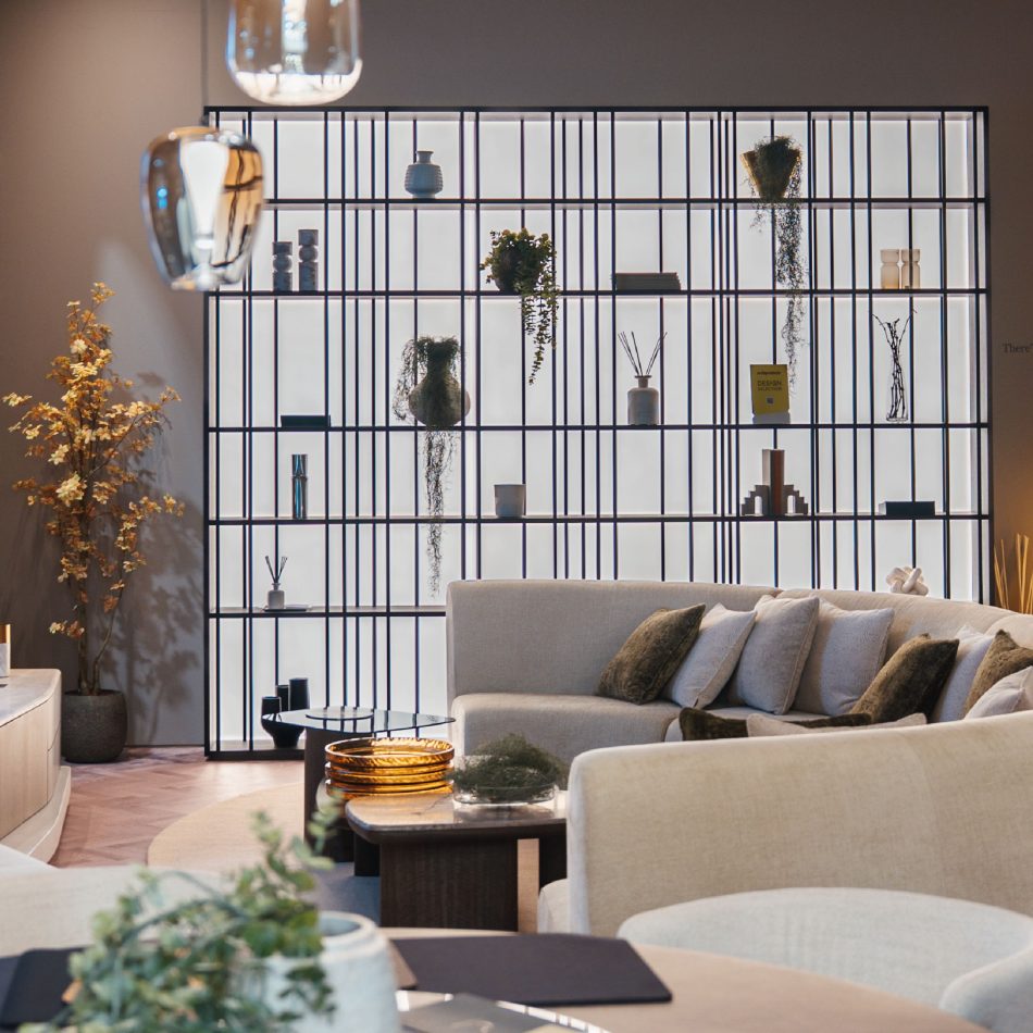 The living and dining area of the stand features a curved beige sofa facing a central coffee table, framed by the illuminated Buren Bookcase displaying ceramics, books, and greenery. Soft lighting and layered materials create an inviting atmosphere shaped by luxury home furniture and warm living room design.