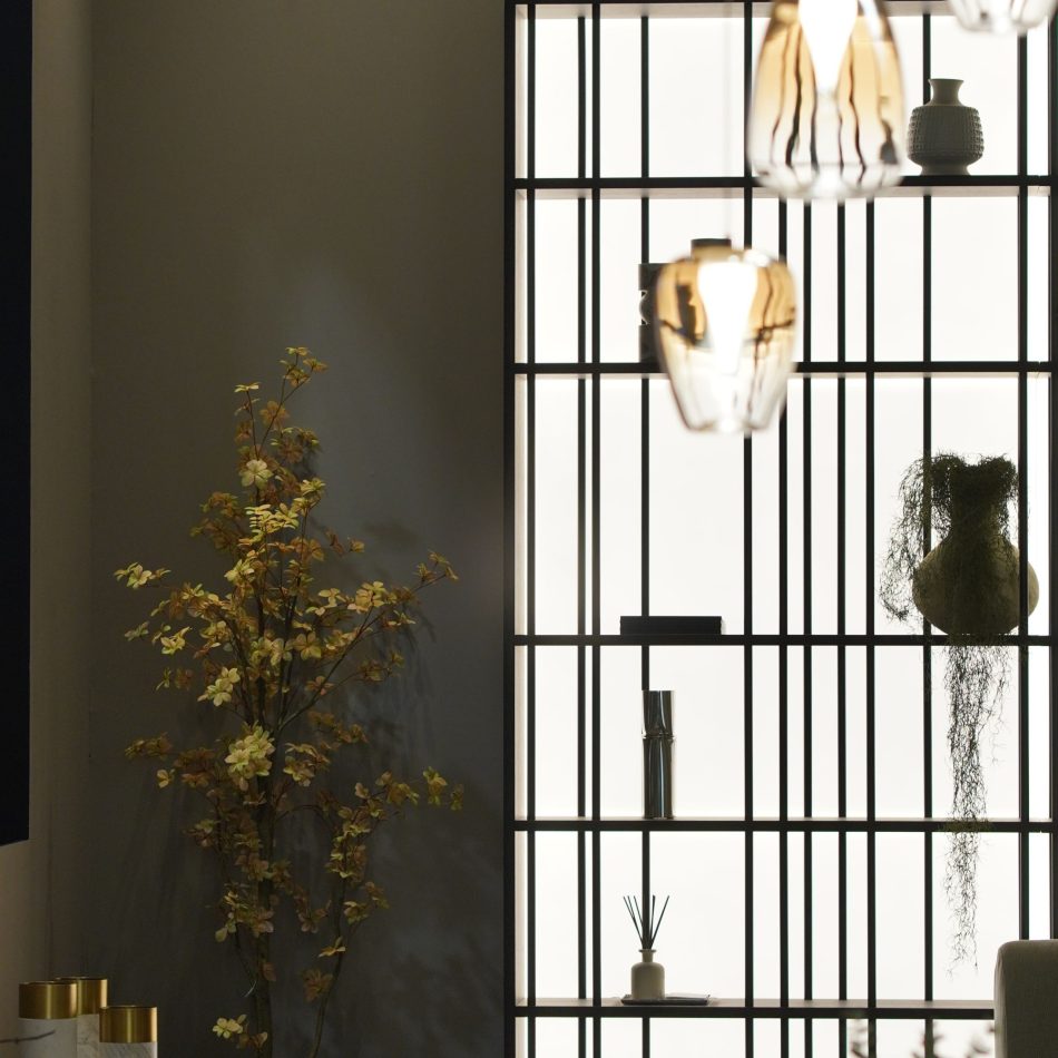 A close view of the illuminated Buren Bookcase reveals a grid of dark metal shelves displaying ceramics, greenery, and textured vases. The warm backlighting and layered objects highlight refined design details and contemporary living room furniture, shaping a sophisticated visual composition.