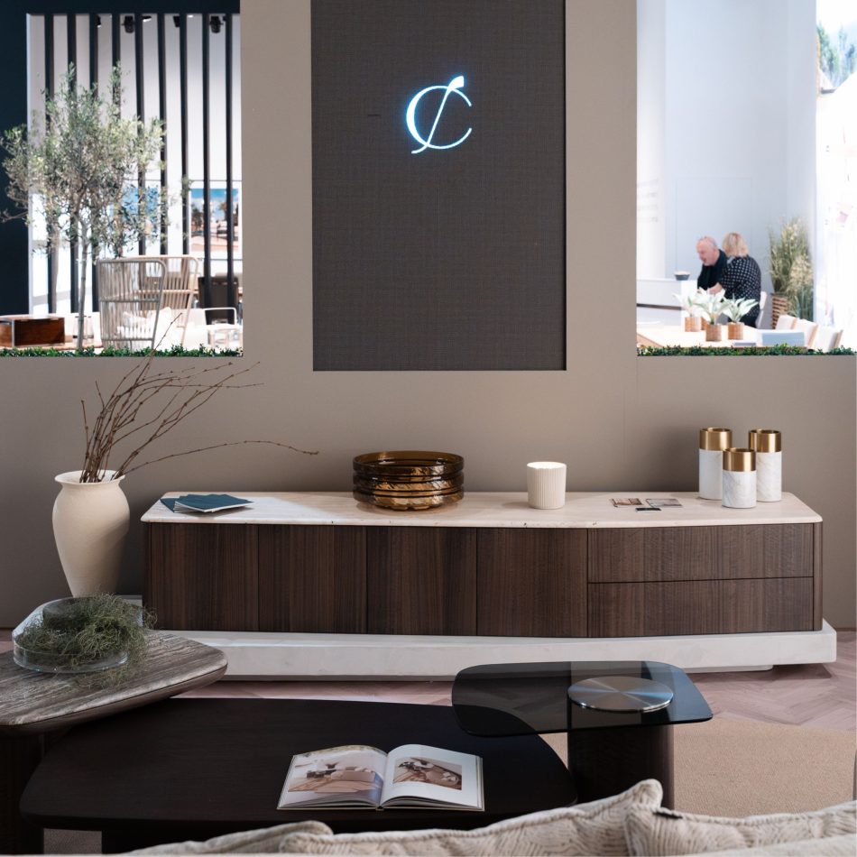 A long Stone TV Cabinet with a marble top and warm wooden structure anchors the space, styled with a white vase, amber glass bowls, candles, and sculptural décor. In front, Dolomites coffee tables hold an open book and a glass bowl with greenery, while glimpses of the adjacent lounge appear through the wall openings. The composition reflects refined living room furniture and contemporary luxury home furniture, shaped by expressive design details.
