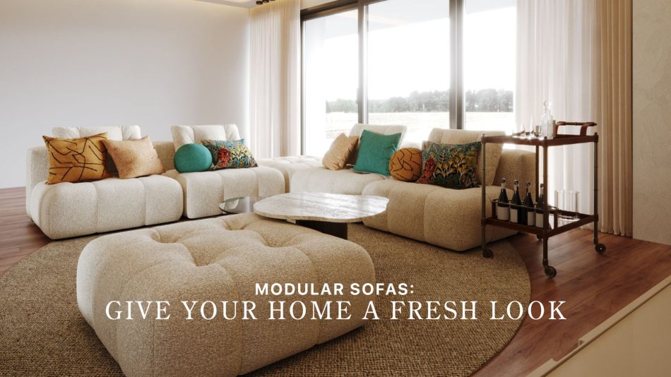 The Antónia Sofa is arranged in a spacious living room as a soft, modular composition in warm neutral tones, styled with colourful cushions that introduce vibrancy and comfort. A matching ottoman and a round stone‑top coffee table anchor the seating area, while a wooden bar cart and floor‑to‑ceiling windows create an inviting, light‑filled atmosphere. The layout highlights the adaptability of modular sofas, showing how thoughtful sofa design can refresh a home with flexible arrangements that evolve over time. Natural textures, layered materials and open views reinforce the harmony and ease characteristic of contemporary furniture.