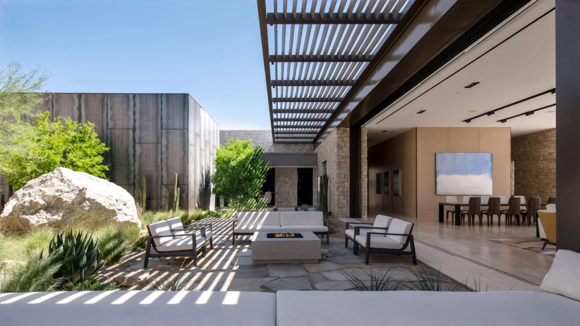 An open-air lounge with white upholstery and dark wood frames surrounds a stone fire pit beneath a slatted pergola, framed by native cacti and sculptural boulders. The fully open glass window reveals a bespoke dining room inside, creating a fluid and effortless living room architecture that embodies minimalist design at its finest.