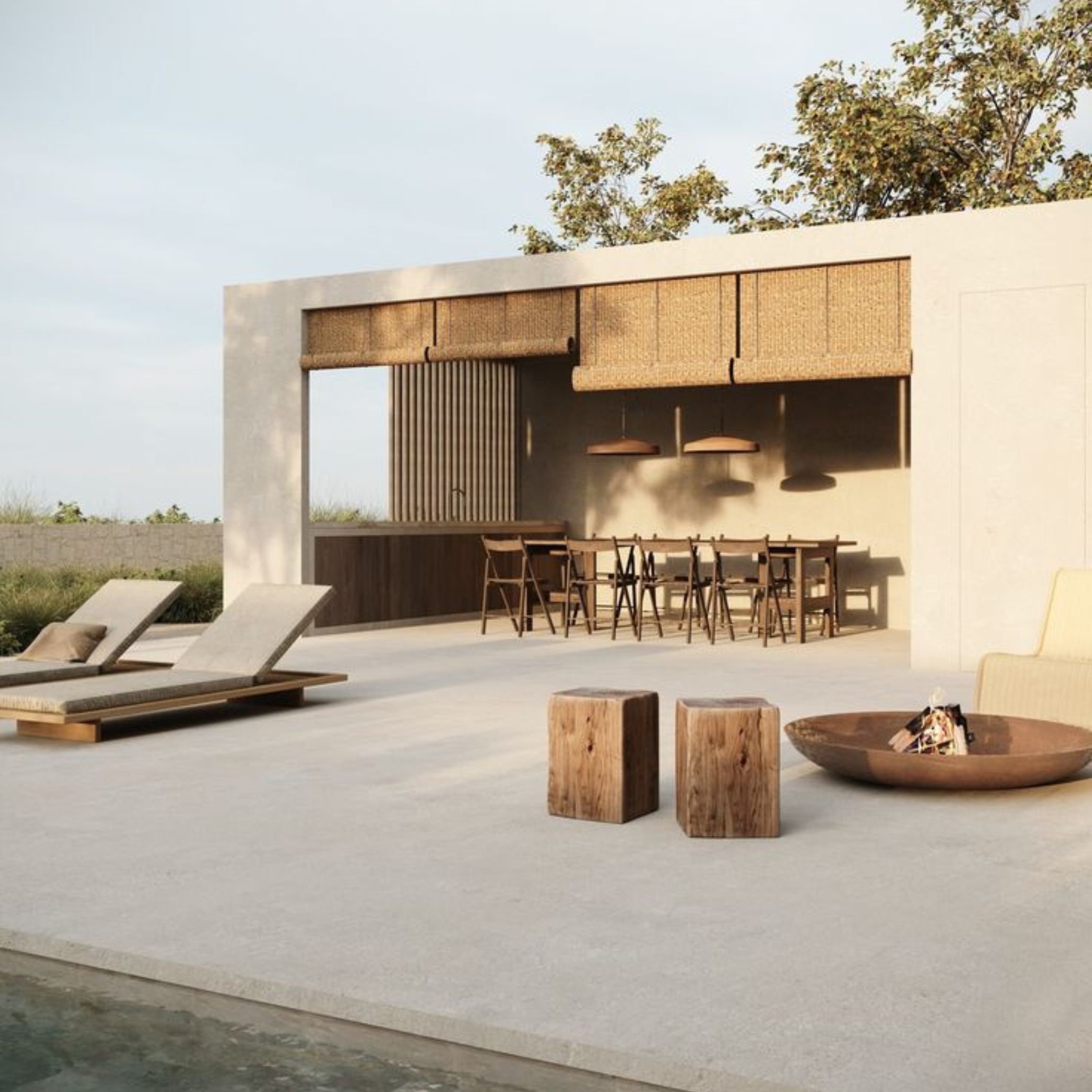 A modern alfresco pool terrace combining a covered outdoor dining area with modern bar chairs along a long wood table, woven shade panels and pendant lighting. Raw solid wood stump side tables and a sculptural fire bowl anchor the open space, reflecting an understated minimalist design where every element is purposeful and connected to the surrounding landscape.