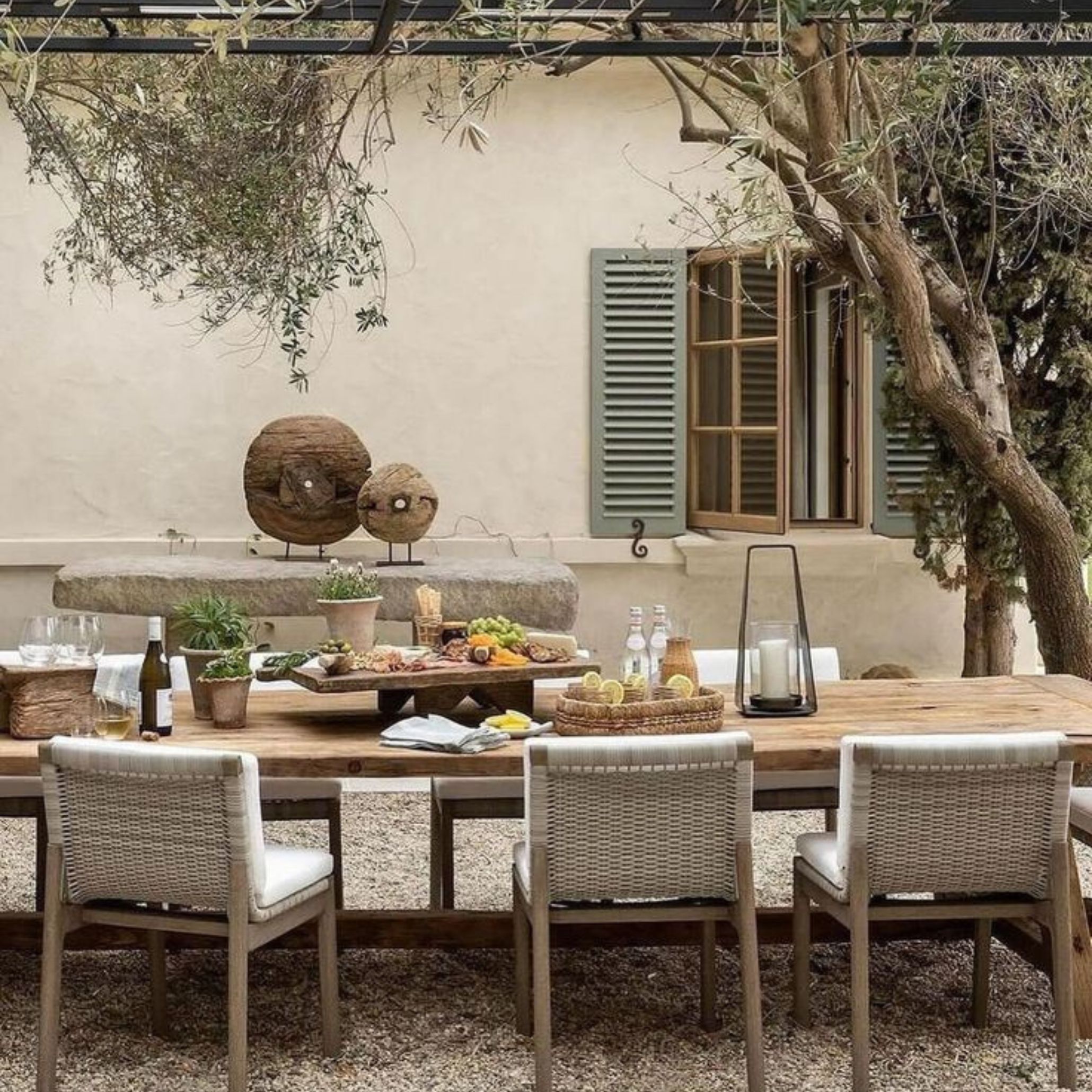 A mediterranean alfresco area outdoors, this Mediterranean courtyard captures the true essence of alfresco living around a long solid wood dining table surrounded by woven restaurant dining chairs with white cushions. Olive trees, a rustic stone bench and an antique plaster facade create a natural backdrop, while the restaurant table setup is dressed with fresh food, candles and organic decorative objects.