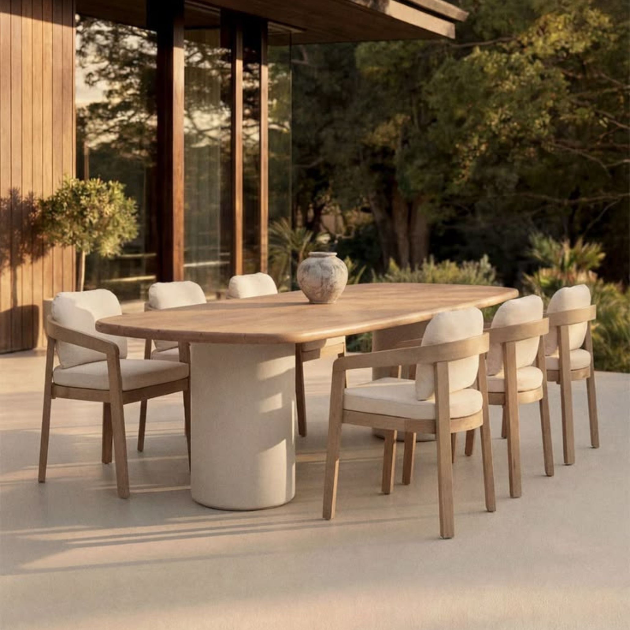 An alfresco modern dining table with an oval solid wood top resting on a cylindrical concrete pedestal, set on a polished terrace beside a timber and glass facade. Eight contemporary dining table chairs in natural wood with cream upholstery surround the table, with a single ceramic vessel as the only decoration. A lush green backdrop completes this refined luxury dining table set designed for outdoor living.