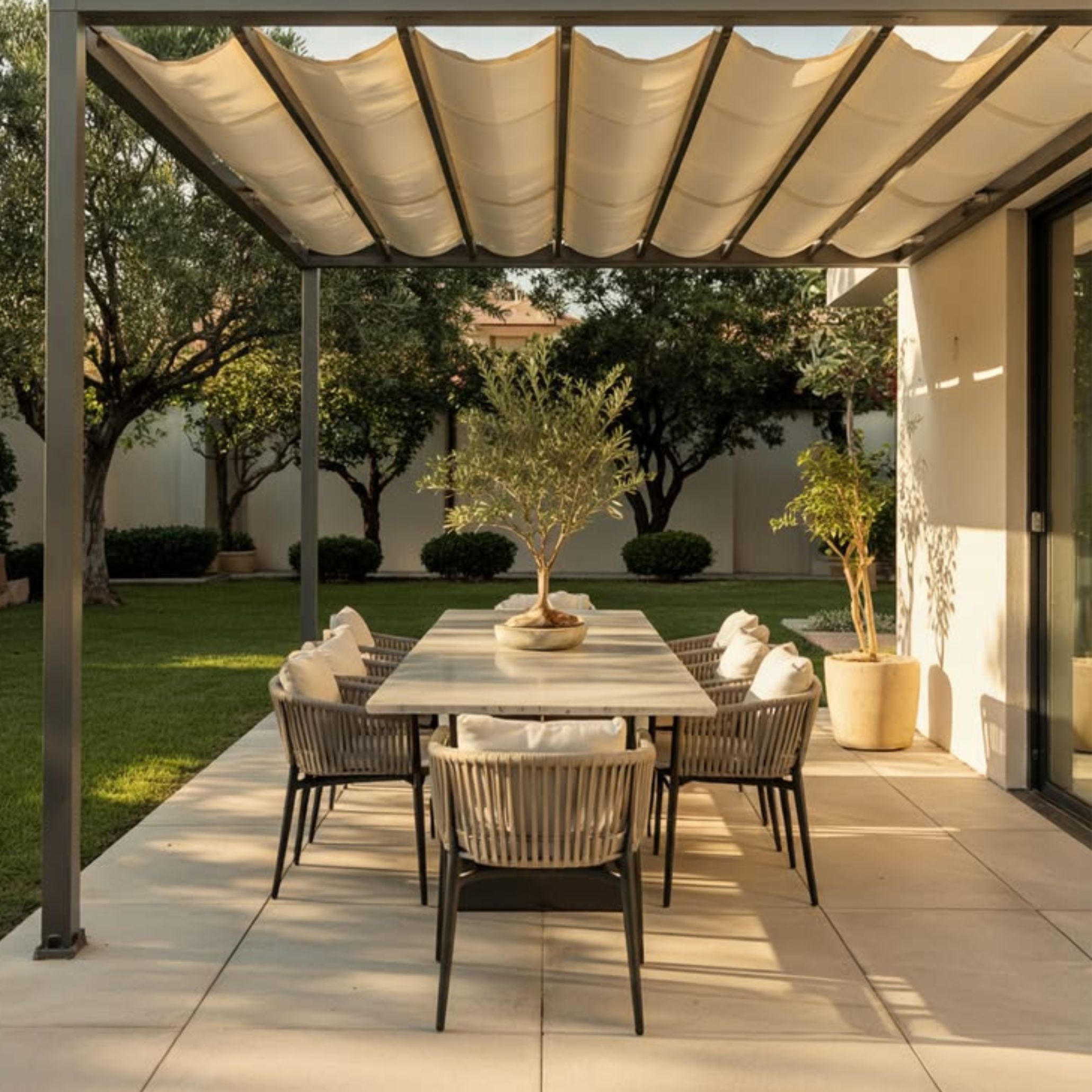 A modern alfresco dining setup beneath a pergola, where a stone-top modern dining table is surrounded by rope-weave dining chairs with white cushioned upholstery. A potted olive tree serves as a natural centrepiece, while the open garden with mature trees provides a lush backdrop. This bespoke dining area brought outdoors captures how shade and natural light work together to shape the alfresco experience throughout the day.
