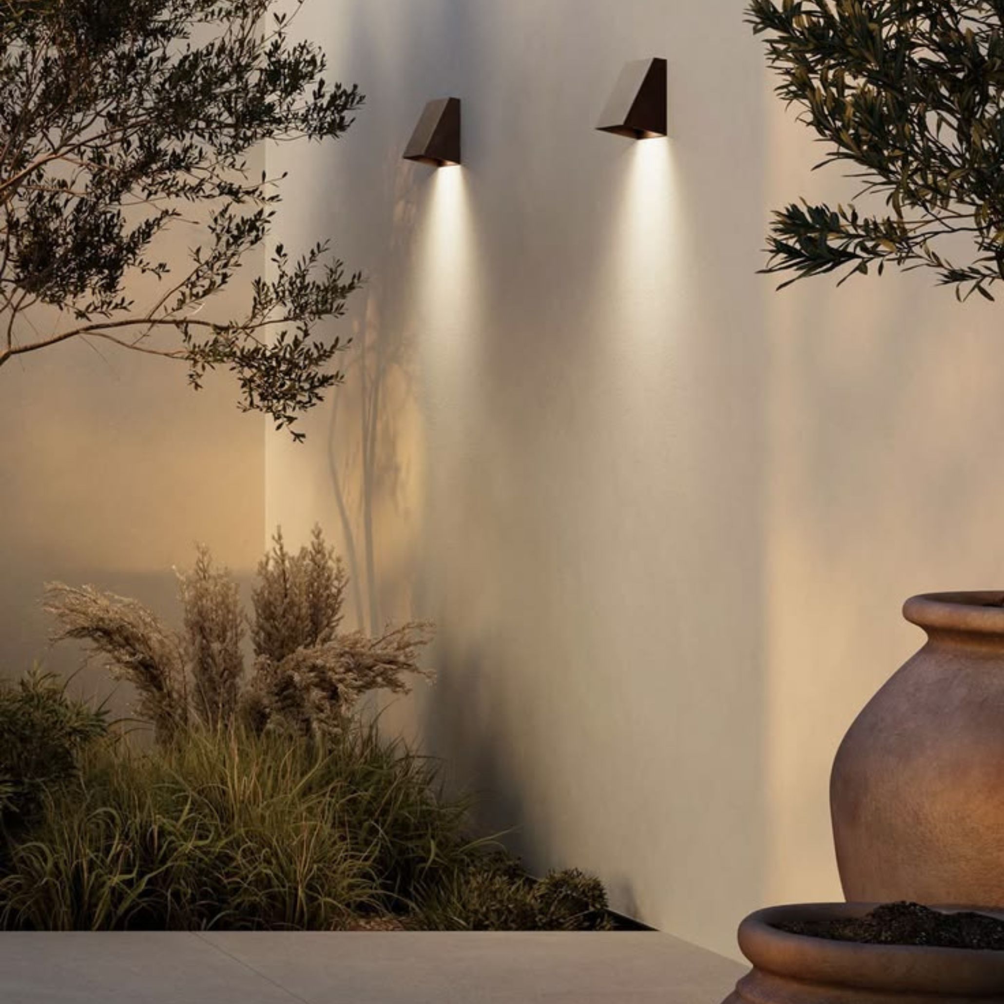 Two angular metal wall sconces cast warm directional light down a white rendered wall, creating a soft and intimate alfresco atmosphere after dark. Ornamental grasses, olive tree branches and a large ceramic vessel compose a layered natural scene that embodies interior design inspiration for outdoor spaces. This image captures how considered lighting transforms an alfresco garden into an inviting setting well into the evening, reflecting minimalist design with carefully chosen decorative details.