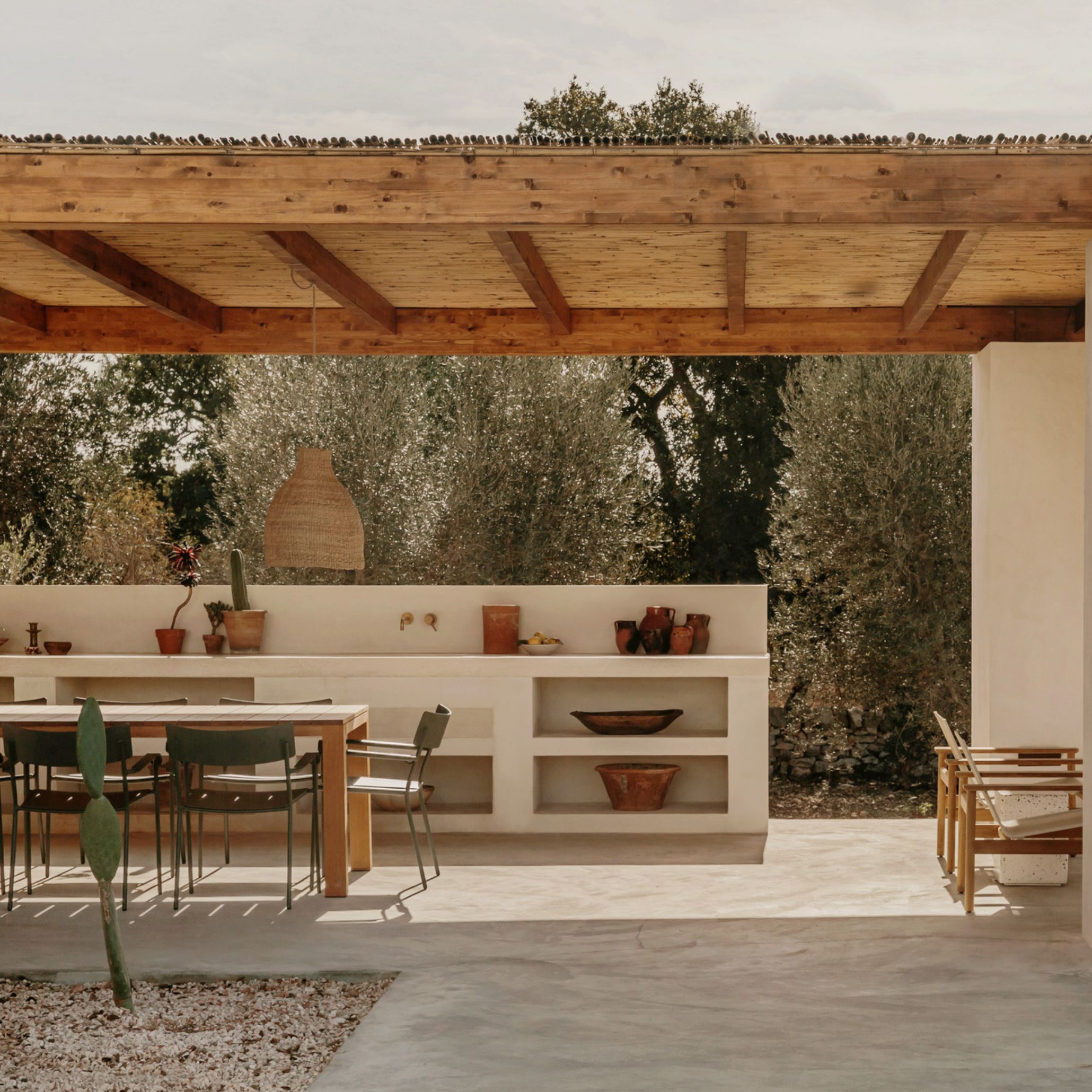 A rustic and modern alfresco space beneath a solid wood beam pergola, combining an outdoor kitchen built in rendered plaster with open shelving displaying terracotta ceramics, alongside a modern dining table with dark metal chairs. The kitchen inspiration extends naturally outdoors here, with brass tap fittings, handcrafted pottery and an olive tree grove as backdrop, embodying the Italian principle that living room architecture and daily life flow freely between shelter and landscape.