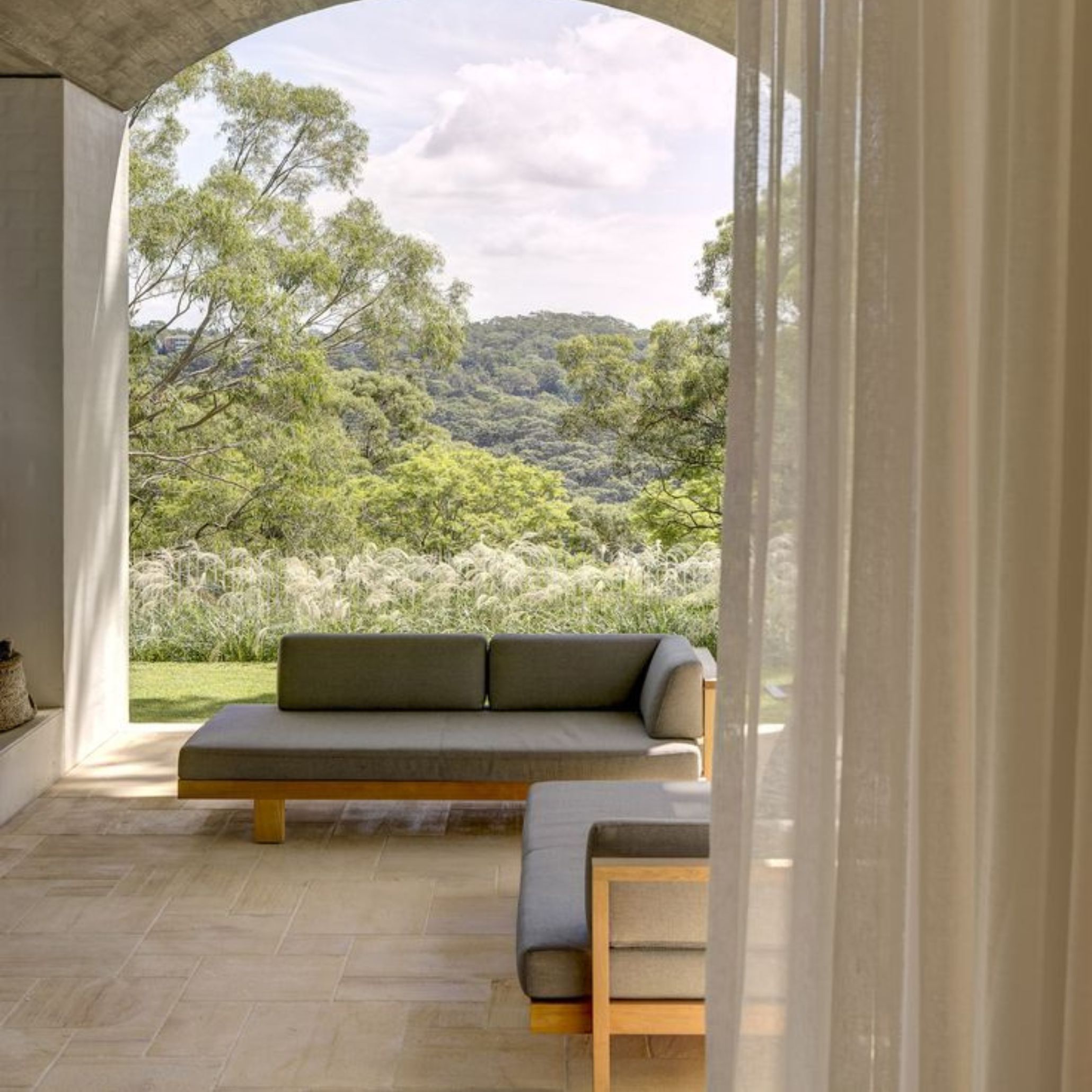 An alfresco covered terrace framed by a wide stone arch and sheer linen curtains, where a low sectional sofa with grey upholstery faces an expansive view of trees and rolling hills. The bespoke living room of this outdoor space, with its solid wood frame furniture and stone tile flooring, captures the Italian ideal of indoor-outdoor flow, where a single seating arrangement becomes a quiet invitation to move freely between both worlds.