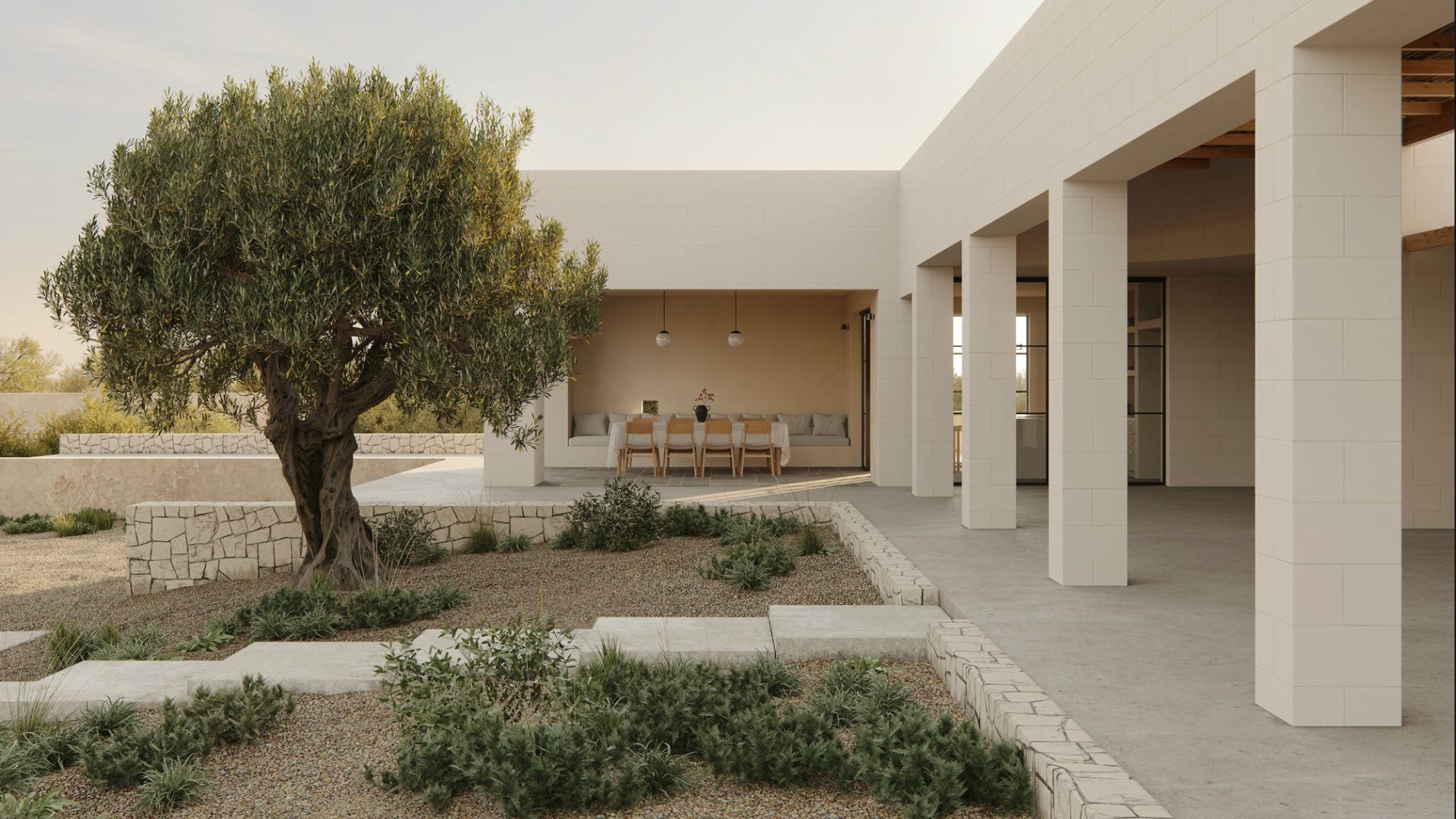 A whitewashed Comporta house wraps around a gravel courtyard anchored by a mature olive tree and low stone-edged planters. Through the open covered terrace, a bespoke dining room with wood chairs and pendant lighting is visible, reflecting the minimalist design principles that define Comporta house decoration. The raw stone retaining walls and drought-resistant planting capture the interior design inspiration of Portugal's Alentejo landscape, where luxury home furniture and natural materials coexist with effortless restraint.