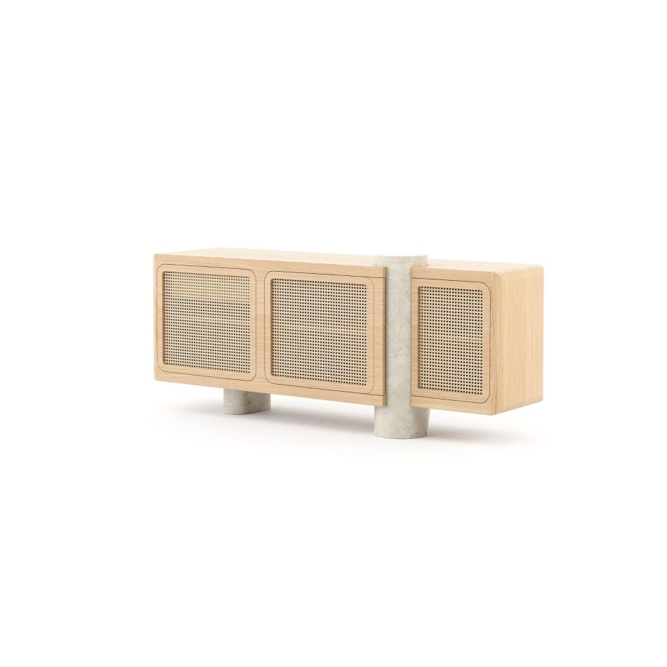 The Antónia Sideboard, a modern sideboard crafted in natural oak wood with a warm, light grain finish. The front reveals three door panels with a fine woven cane surface, while a smooth, rounded travertine marble column rises vertically through the case between the second and third doors, with a matching cylindrical marble leg visible at the base. A elegant choice of luxury dining room furniture for spaces that mixes texture, wood, and stone.
