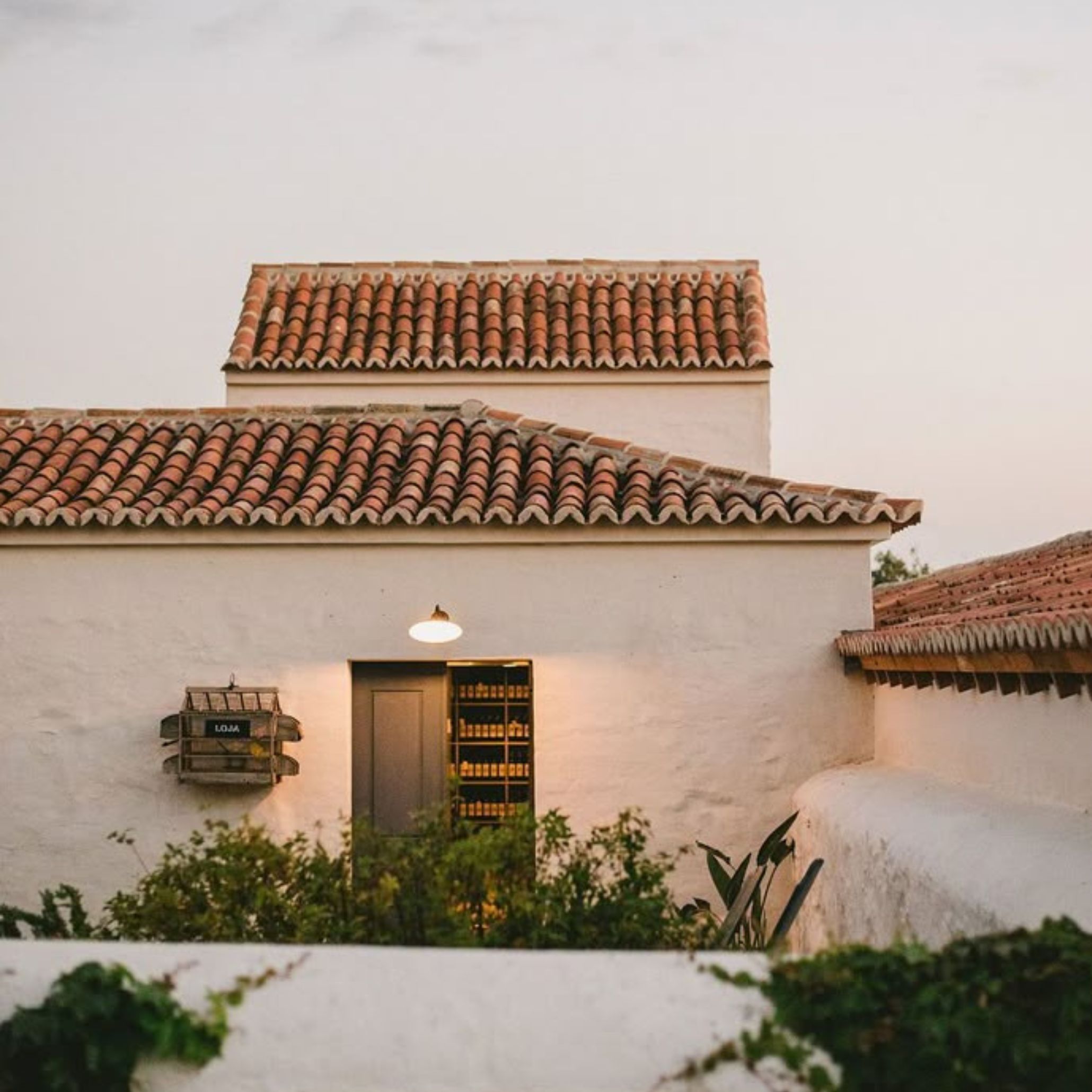 A whitewashed Comporta house at dusk with layered terracotta tile roofs and a warm-lit doorway opening onto shelved interior storage. The raw textured walls, climbing plants and handmade wood design signage reflect the minimalist design and material honesty that define the Comporta aesthetic, where architecture serves as interior design inspiration rooted in handmade furniture traditions and the Alentejo landscape.