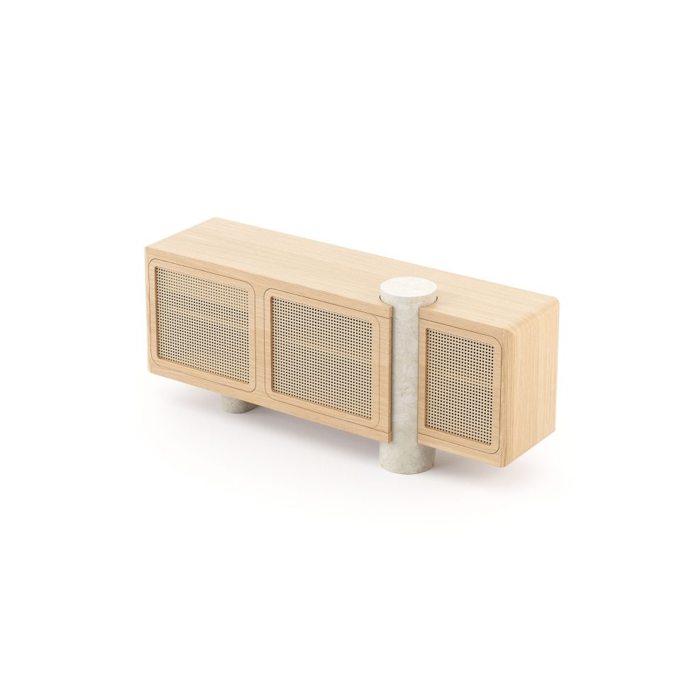 The Antónia Sideboard showing the natural oak wood casing and its three woven straw door fronts in close detail, with the wicker pattern catching the light. The travertine marble post is visible running through the structure, capped with a flat circular marble disc at the top, contrasting the soft stone texture against the wood grain. A sculptural anchor piece for living room furniture collections.