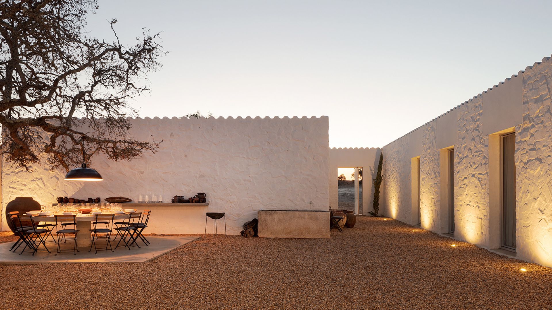 An outdoor dining setup at dusk within a whitewashed stone courtyard, where a long dining room tables in pale stone is surrounded by dark folding chairs beneath a single pendant light. Terracotta vessels and handwoven objects line a built-in ledge against the textured wall, while warm uplighting traces the gravel path toward an open archway. The sandy tones and raw wood texture of this bespoke dining room reflect the earthy Comporta colour palette drawn directly from the Alentejo landscape, grounding the space in interior design inspiration rooted in Portuguese tradition.