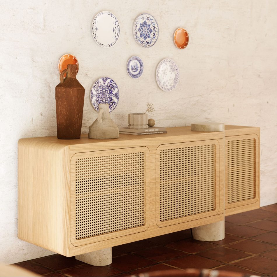 The Antónia Sideboard in natural oak wood with straw doors and a rounded travertine marble leg, set against a rustic whitewashed wall in a Comporta-style home. The piece anchors the room, styled with terracotta vases, hand-thrown ceramics, and a curated wall of Portuguese decorative plates above, with a terracotta tiled floor completing the warm, earthy palette. A grounding piece of interior design inspiration for relaxed coastal interiors.