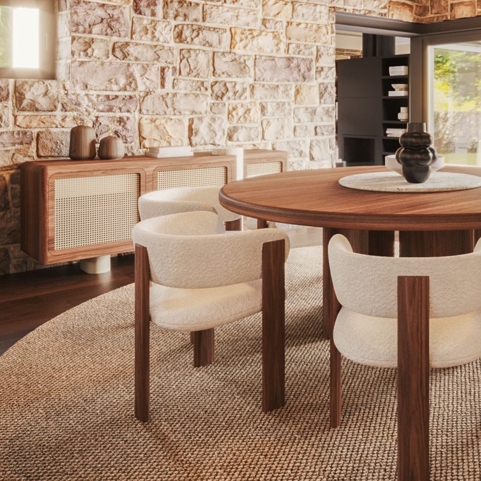 The Antónia Sideboard in a deep walnut wood finish, wall-mounted against a textured natural stone wall in a dining room setting. Its three woven cane door panels and travertine marble leg are visible just behind a round walnut dining table surrounded by cream boucle armchairs, with a woven jute rug underfoot. The sideboard's grounded, sculptural presence makes it a fitting centerpiece for bespoke dining room schemes.