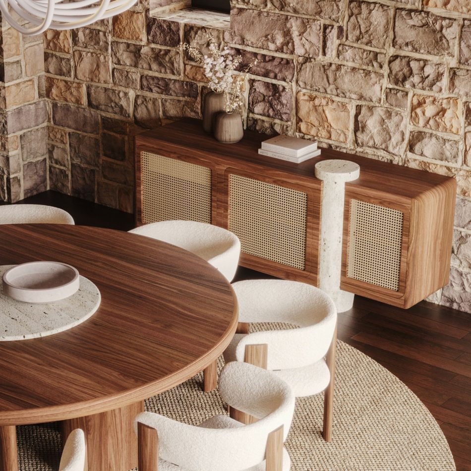 The Antónia Sideboard in walnut wood, mounted against a rough-cut stone wall and styled with ribbed ceramic vases and stacked books on top. The woven cane doors and travertine marble column are shown in detail beside a round wooden dining table and upholstered boucle chairs, highlighting the sideboard's role as a refined focal point within the wider room. A study in design details, where wood, cane, and stone come together.