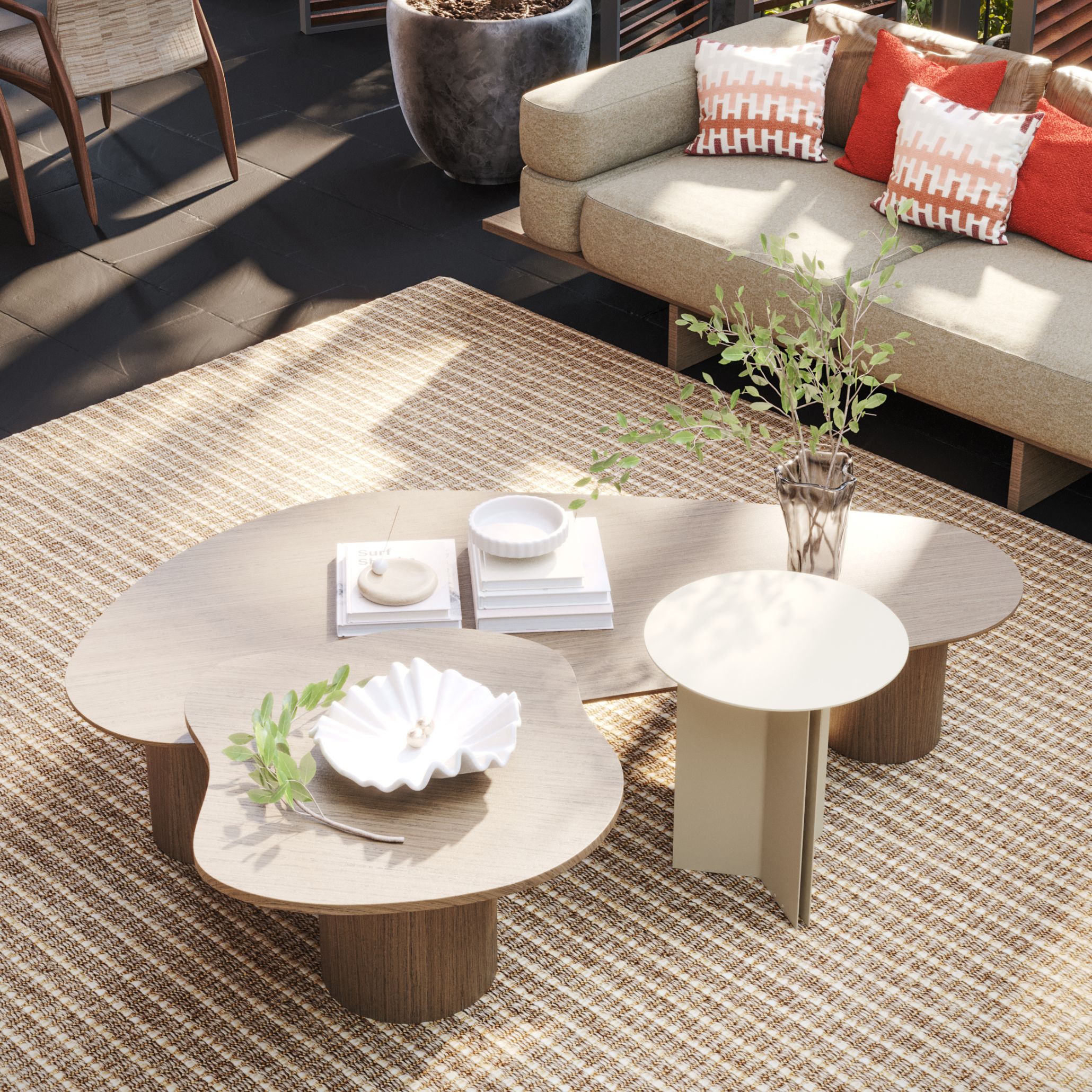 A close-up of the Natur Coffee Table with its organic free-form wood texture top in warm oak, styled with a ceramic shell dish, stacked books and a glass vase, paired with a smaller cream metal side table. The Ripa Sofa with red and patterned pillows is visible behind, the full setting resting on a woven jute-style rug that reflects the minimalist design and natural material language of Comporta outdoor living.
