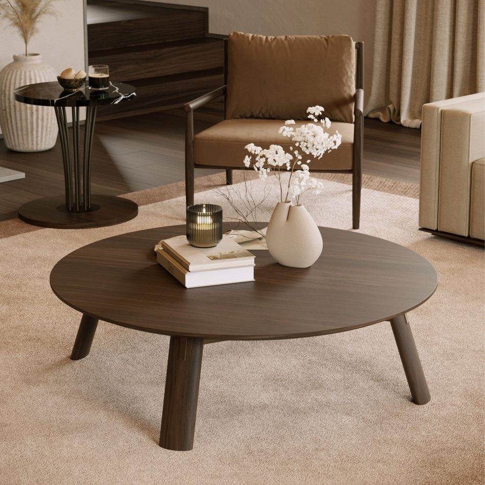 Close view of a layered living room of leather armchairs, a marble side table, and soft drapery, the Landform Coffee Table's round oak top and tapered legs take center stage, dressed with stacked books, a ribbed candle holder, and a sculptural ceramic vase. The honest material and balanced proportion highlight this furniture luxury design as a true luxury coffee table.