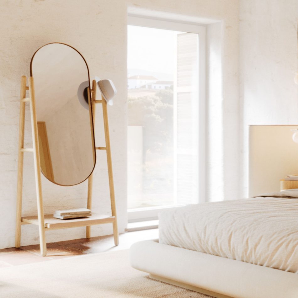 Bright, minimal bedroom corner with the Landform Mirror in natural solid wood A-frame structure positioned beside a floor-to-ceiling window flooding the space with light. The oval brass-framed mirror glass reflects the landscape beyond, while a stacked books shelf at the base adds quiet functionality. A defining bedroom mirror long piece for a bespoke bedroom that values simplicity and interior design inspiration rooted in natural form.