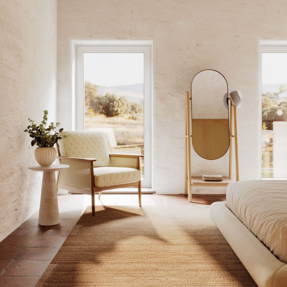 Light-filled Comporta-style bedroom with whitewashed walls, terracotta tiles, and a woven rug, featuring the Landform Mirror in natural solid wood with an aged brass oval frame standing against the wall between two tall windows. A rattan armchair and stone side table complete the scene in the foreground, establishing the mirror as a natural anchor of mirror decor and luxury home furniture within a bespoke bedroom of quiet, considered character.