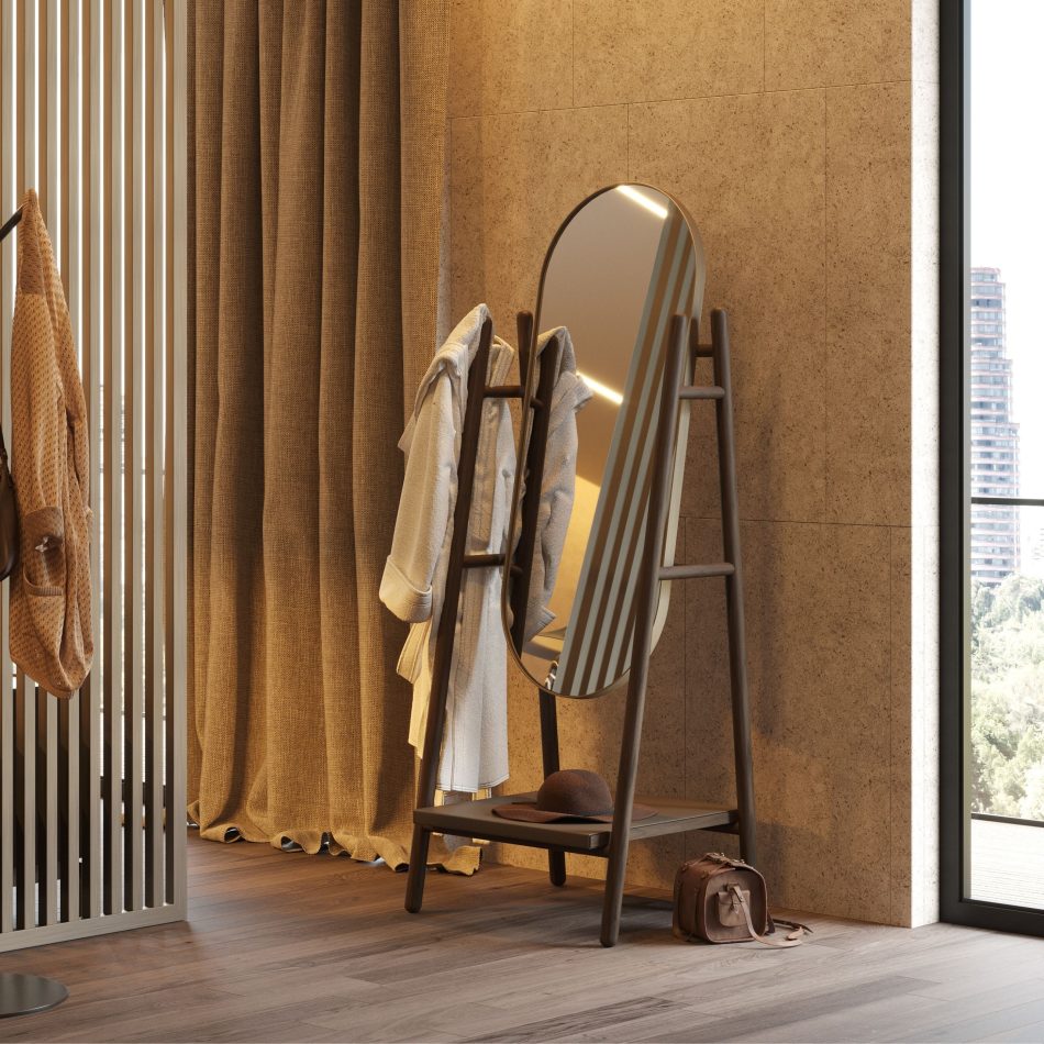 Warm dressing area with cork wall cladding, ochre curtains, and a wood slat partition, featuring the Landform Mirror in dark solid wood with an oval brass frame used as a functional valet stand, with garments draped over the A-frame uprights and a hat resting on the lower leather upholstery shelf. A scene of mirror for interior design that captures the piece's versatility as both mirror decor and everyday companion within a bespoke bedroom or dressing space of genuine warmth and design details.